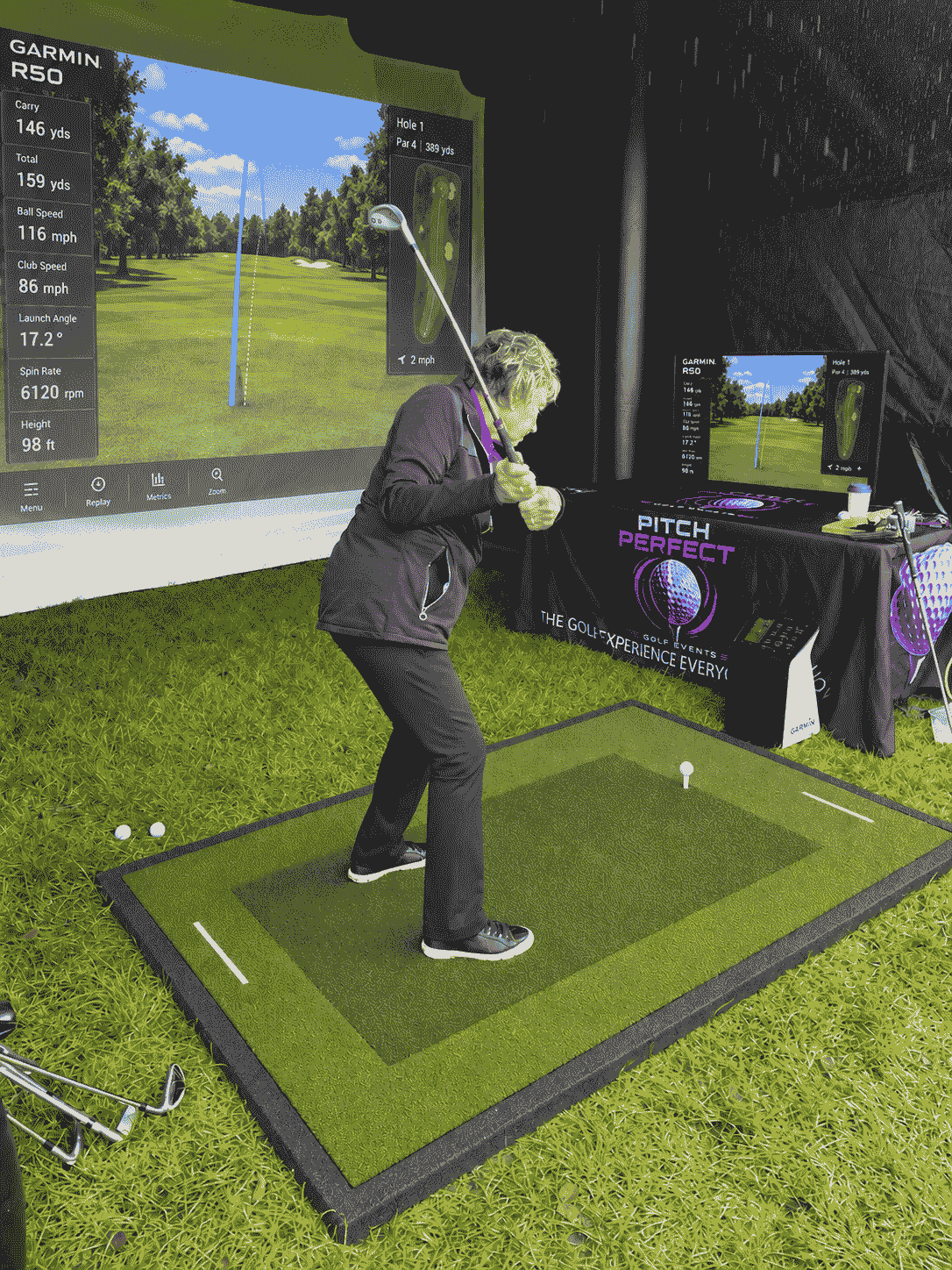 A female golfer swinging a club in front of the impact screen and TV screen which both show the flight of the ball
