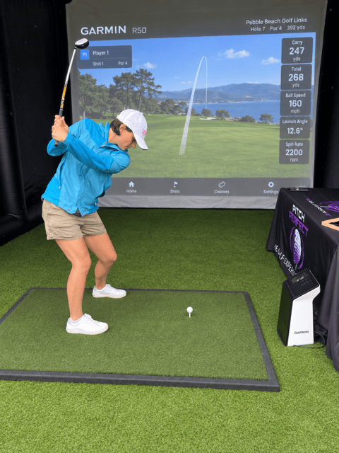A female adult swinging a golf club inside an inflatable golf bay. Also showing an image of a golf course on the impact screen at the back of the bay 