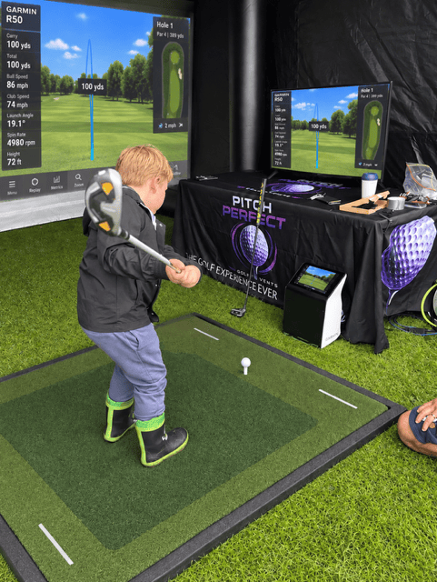 A small male child golfer swinging a club in front of the impact screen and TV screen which both show the flight of the ball