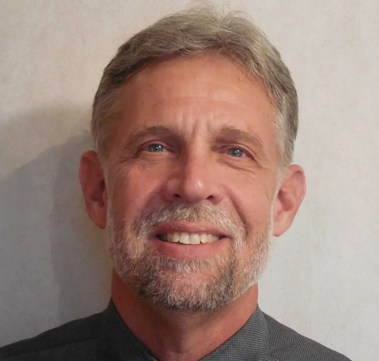A smiling middle-aged man with gray hair and a beard, dressed in a dark shirt, standing against a plain off-white wall.