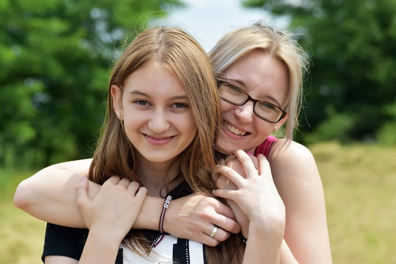 A young woman with long light brown hair and an older woman with blonde hair and glasses smiling outdoors, embracing each other.