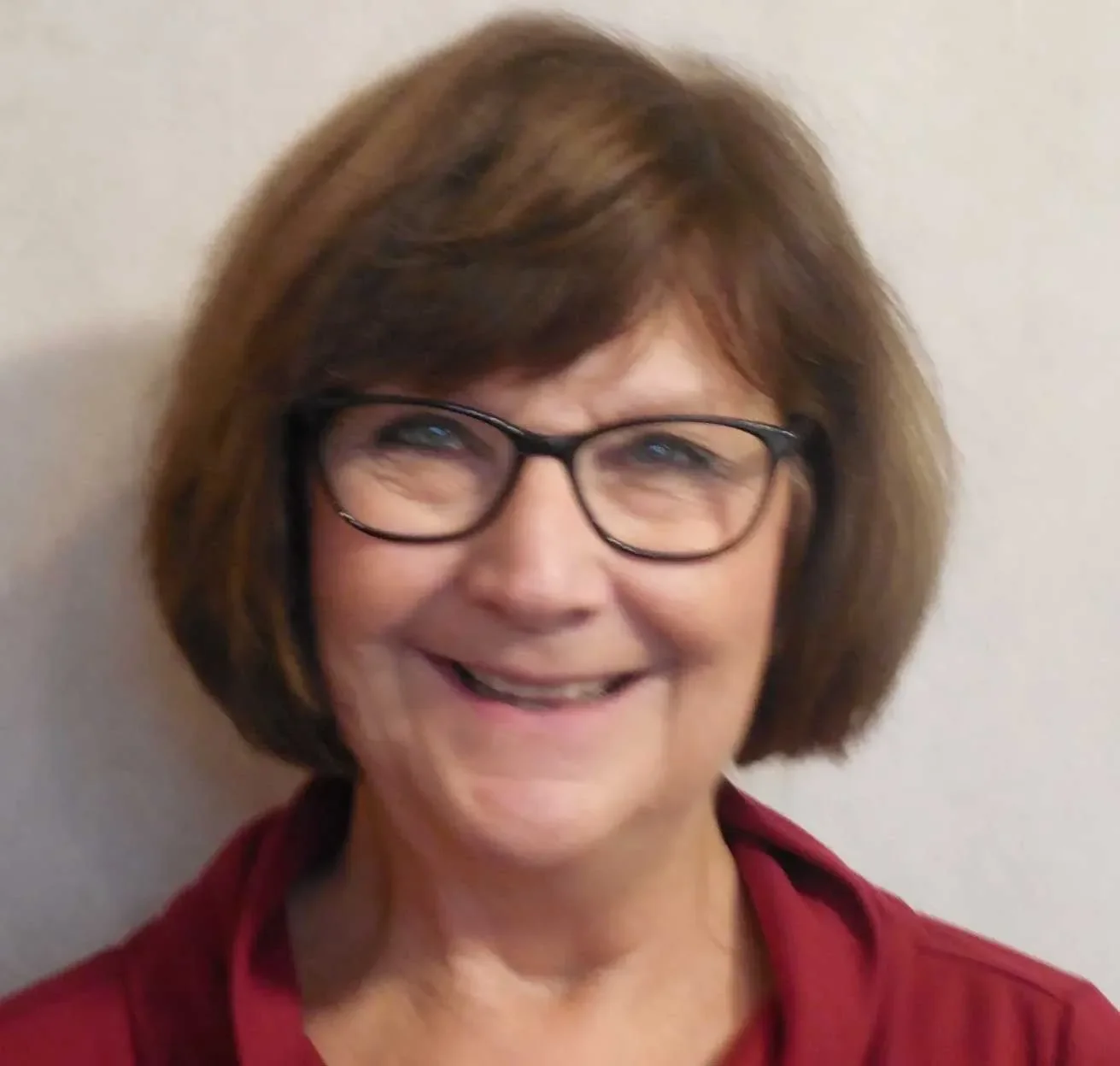 A smiling woman with short brown hair, wearing black glasses and a red top, standing against a plain light-colored background.