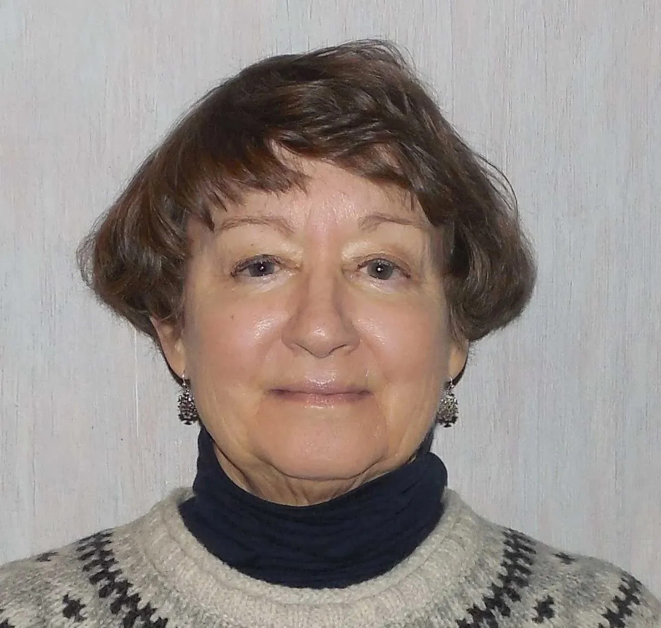 A woman with short brown hair, wearing earrings, a black turtleneck, and a beige sweater with a black pattern, standing against a light-colored wall.