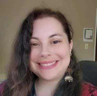 A woman smiling with long, dark, wavy hair, wearing earrings with a decorative element, and a maroon top.