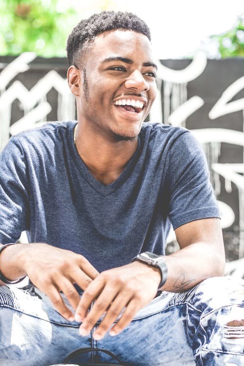 A young man sitting outdoors, smiling, wearing a dark blue t-shirt and ripped jeans, with graffiti art in the background.