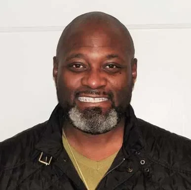 A smiling Black man with a beard and bald head, wearing a black jacket and a gold chain, standing against a plain light background.