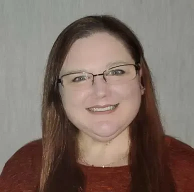 A woman with long brown hair, glasses, and a maroon top, smiling in front of a neutral background.