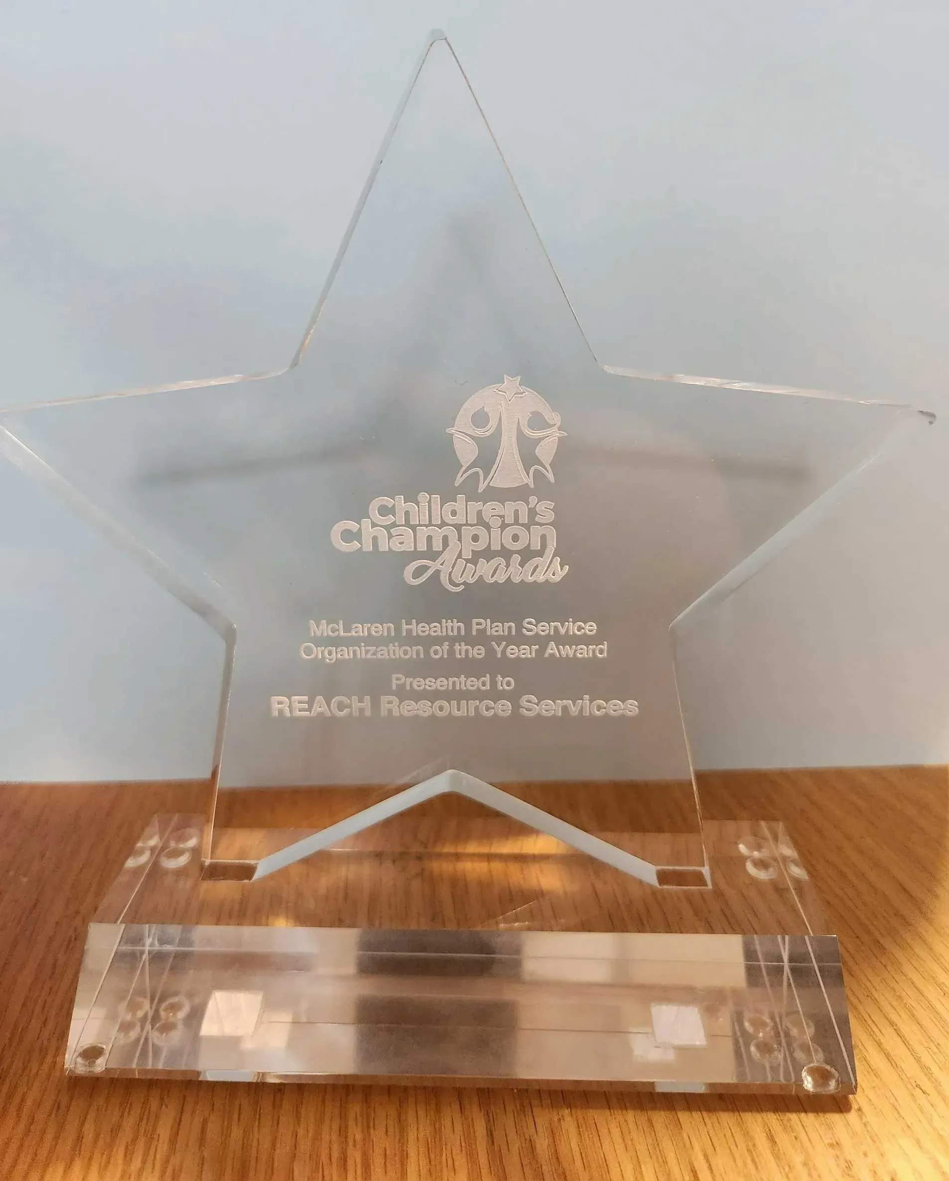 A star-shaped clear acrylic award with engraved text for the Children's Champion Award, presented to Reach Resource Services by McLaren Health Plan Service. The award stands on a wooden surface.