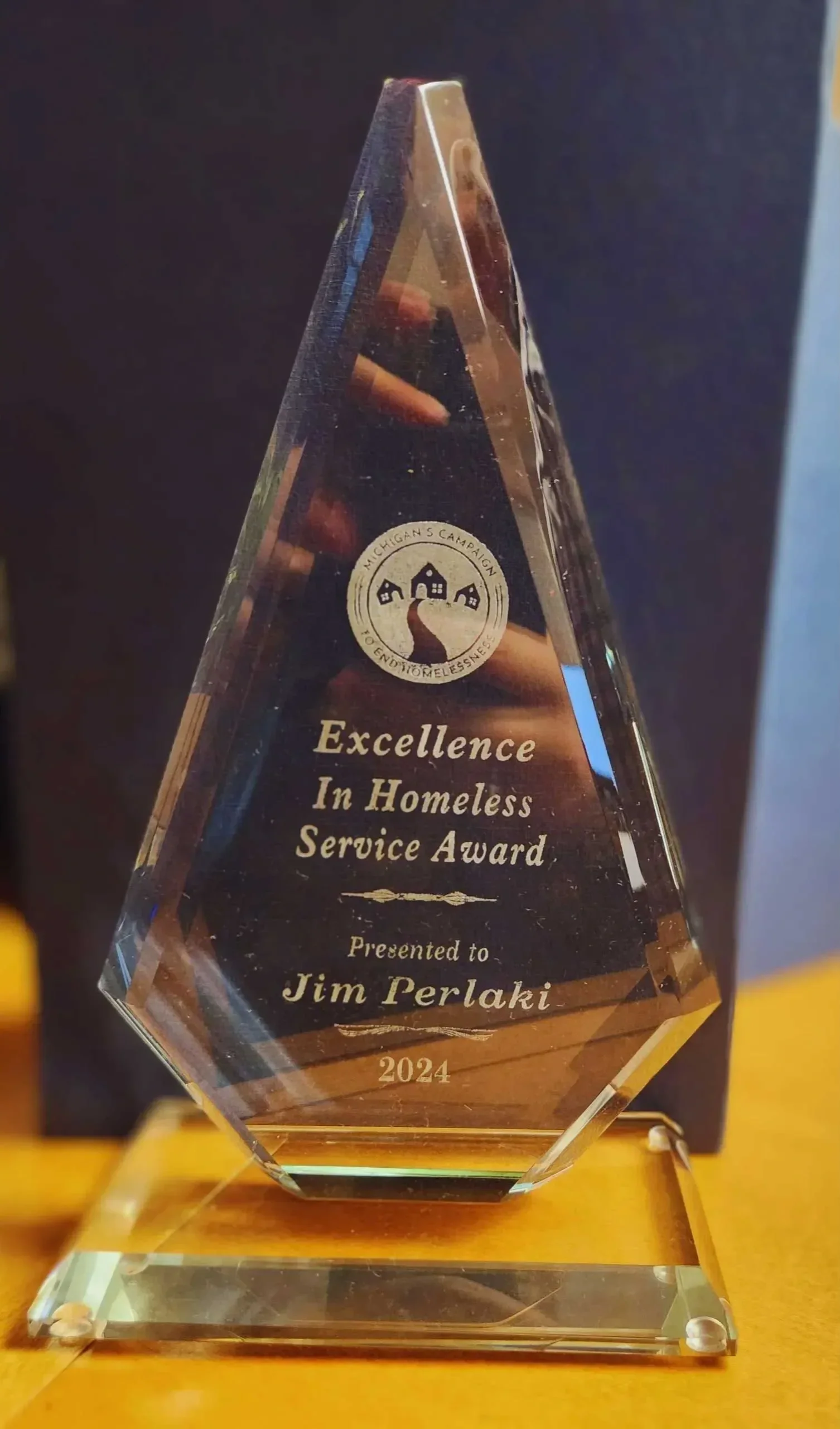A glass award trophy with engraved text that reads: Excellence In Homeless Service Award, presented to Jim Perlak by Michigan's Campaign, 2024.