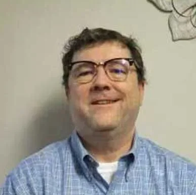 A middle-aged man wearing glasses and a blue checkered shirt, smiling, standing against a plain wall.