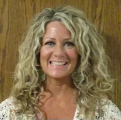 A woman with long, curly blonde hair smiling in front of a wooden wall.