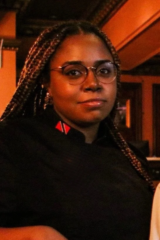 A woman with braided hair, wearing glasses and a black shirt, in a warm, dimly lit indoor setting.