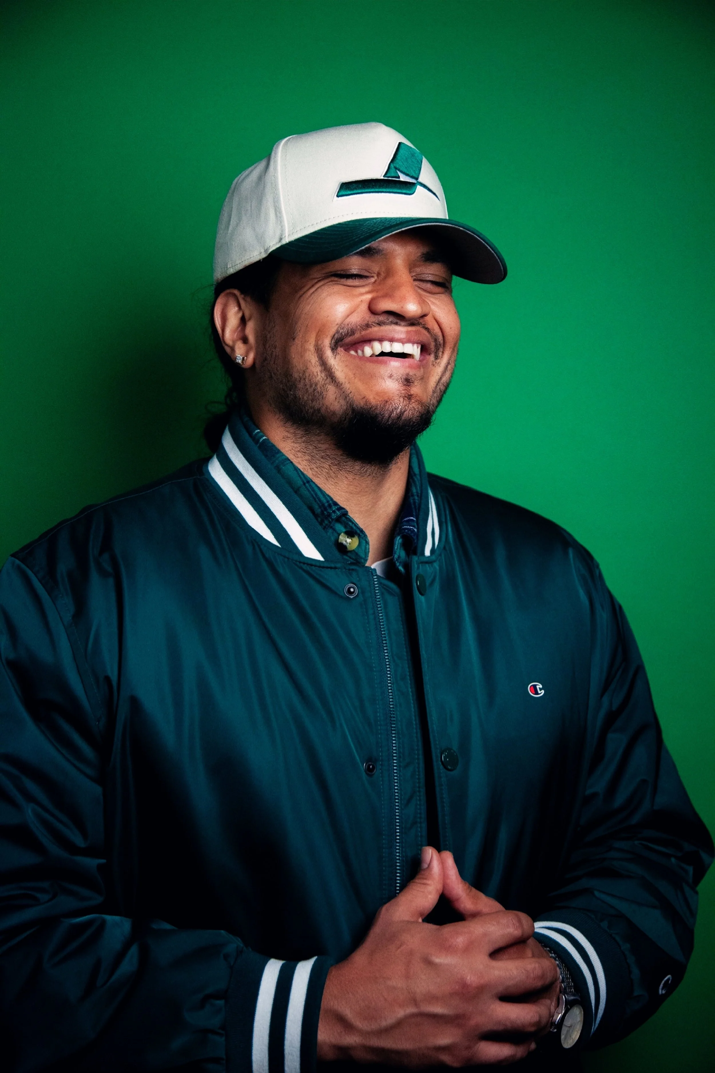 A man smiling wearing a baseball cap and a black jacket with white and teal accents against a green background.