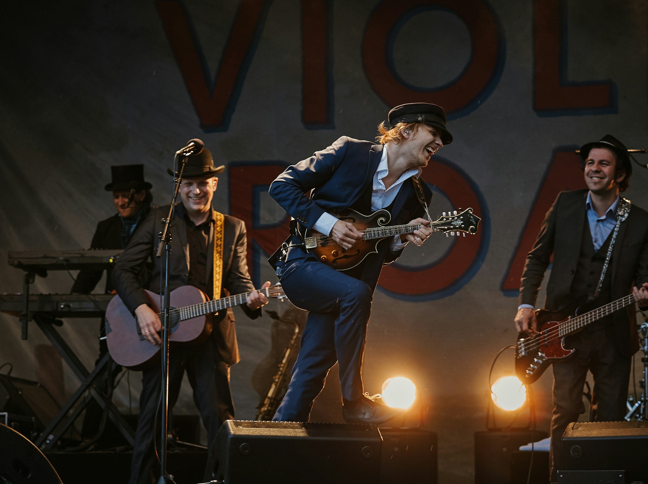 Musicians performing on stage, including a man playing a mandolin, a man playing an acoustic guitar, and others with guitars, under warm lighting with large letters in the background.