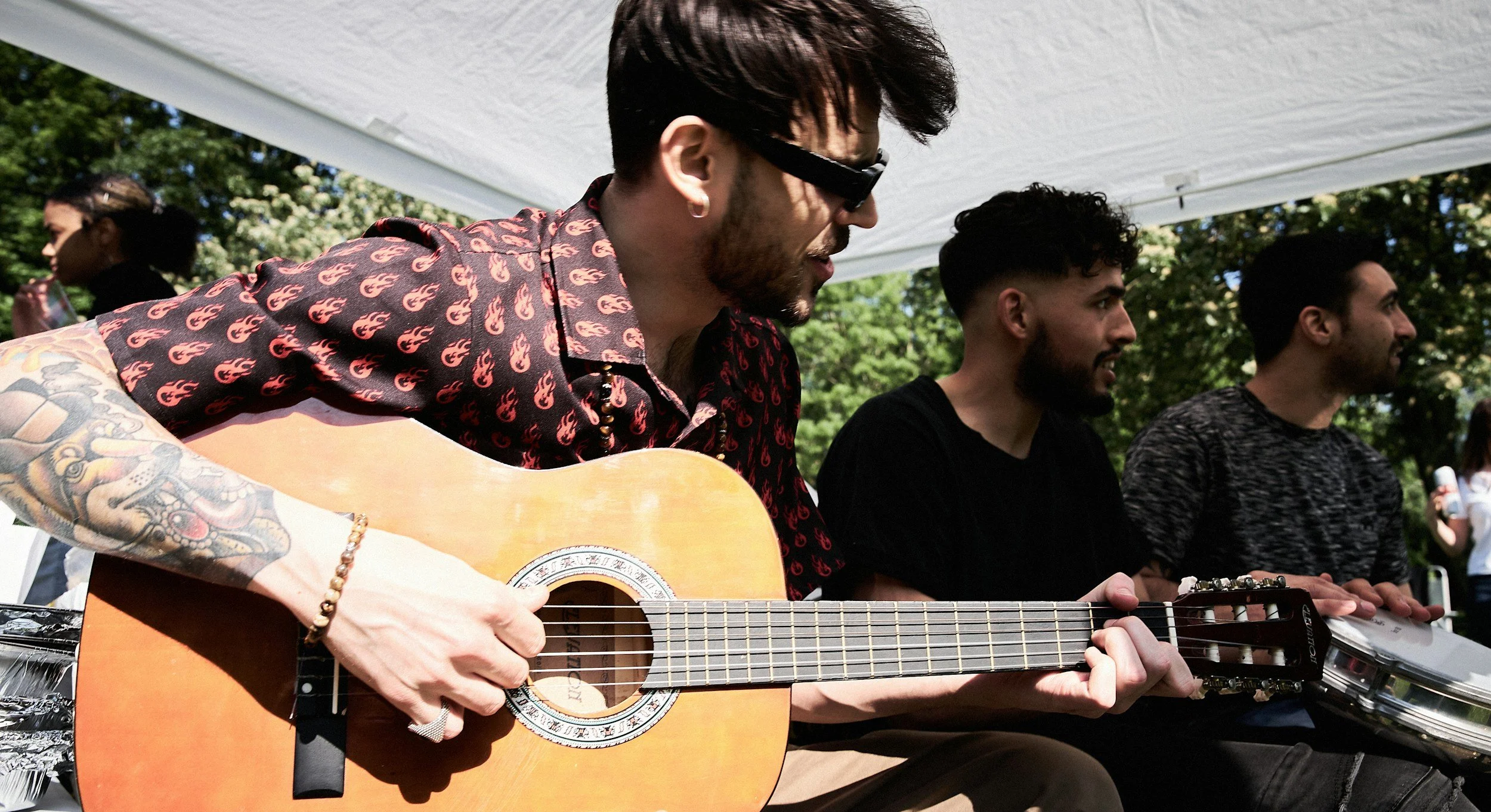 A group of musicians playing guitars and percussion outdoors under a white canopy, with trees and sunlight in the background.