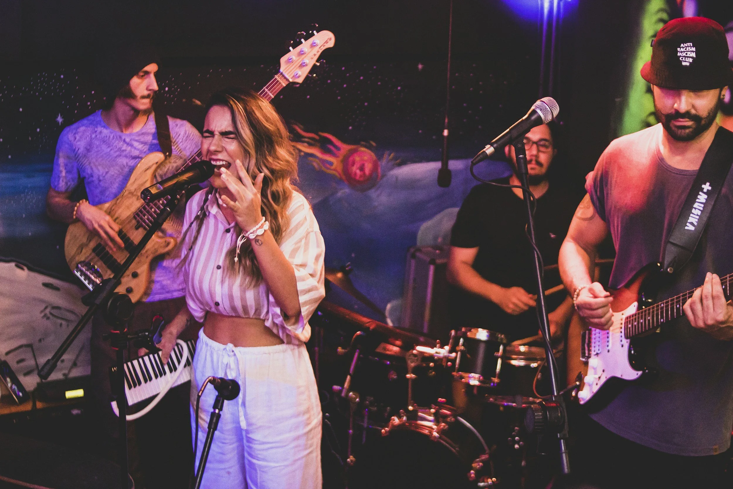 A musical band performing on stage with a starry space-themed backdrop. The band includes a woman singing into a microphone, a man on guitar, and other members playing instruments. The woman has long hair and is wearing a striped, cropped shirt, and the man has a beard and is wearing a bucket hat with text on it.