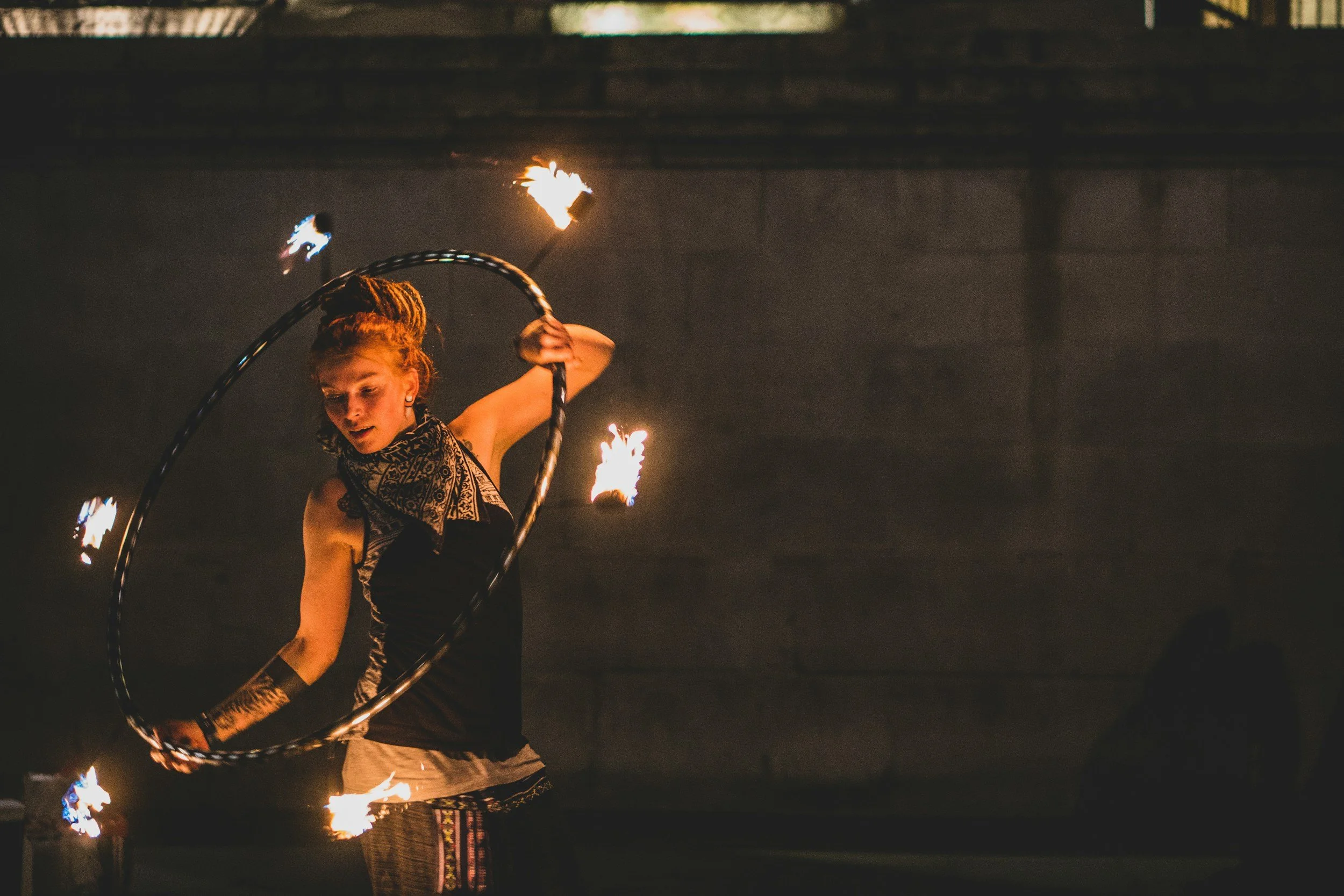 A person with styled hair and tribal face paint performing a fire spinning act at night, holding a lit object creating flames, with a dark background.