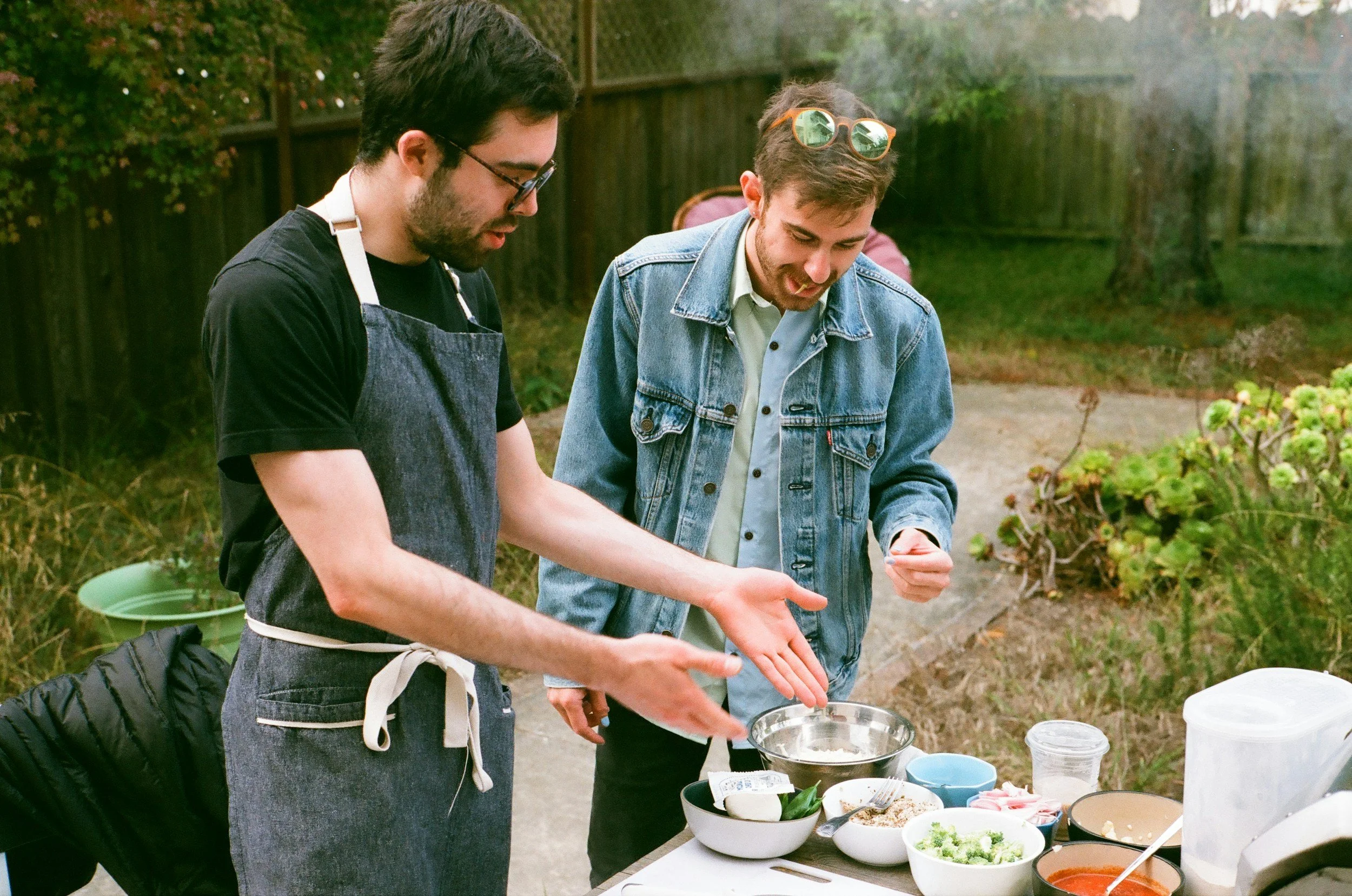 Two young men with glasses, wearing casual clothing and an apron, preparing food outdoors at a table with various bowls and ingredients.