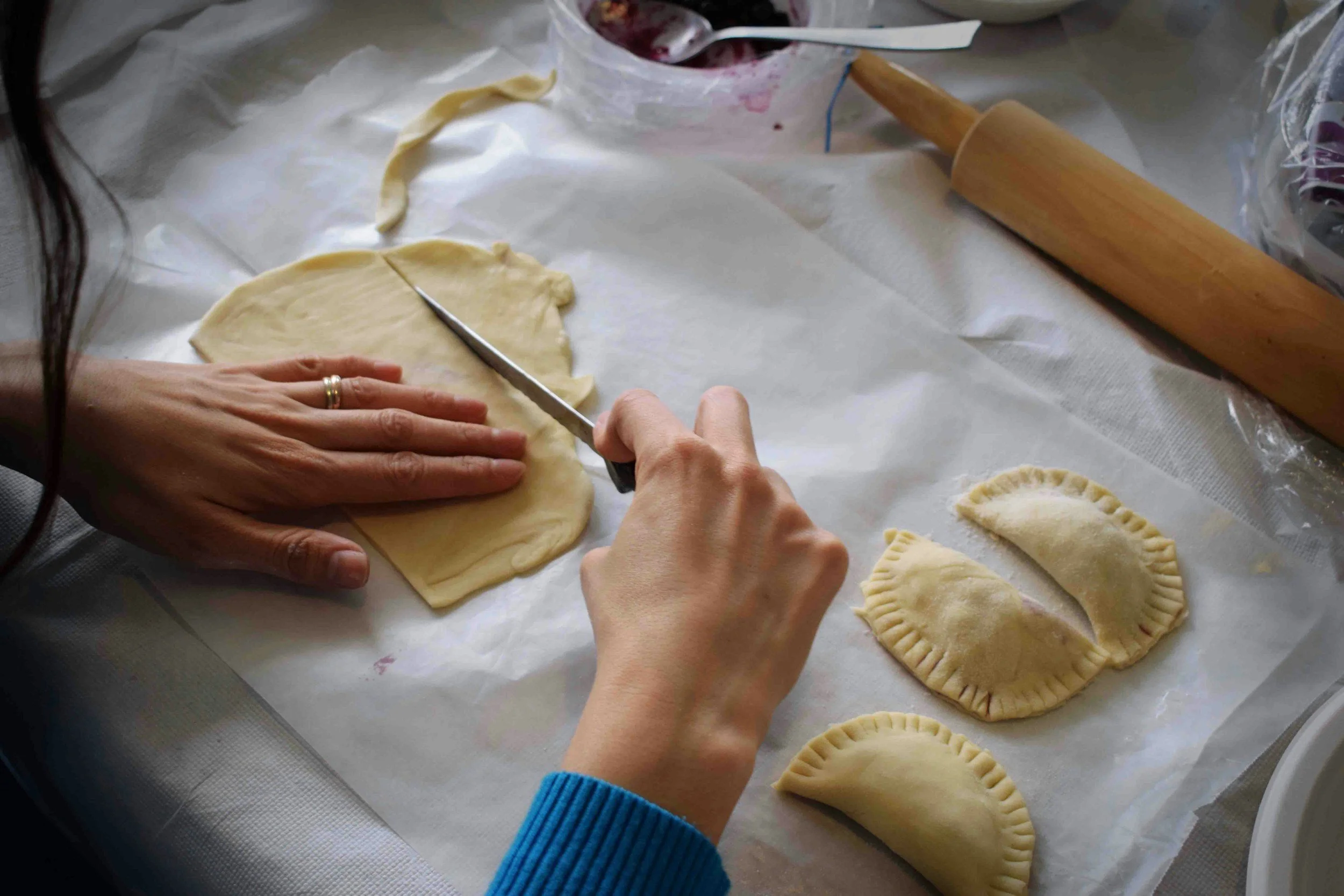 Person cutting rolled-out dough with a knife on a surface covered with parchment paper, surrounded by unbaked pastries, a rolling pin, and a bowl of filling.