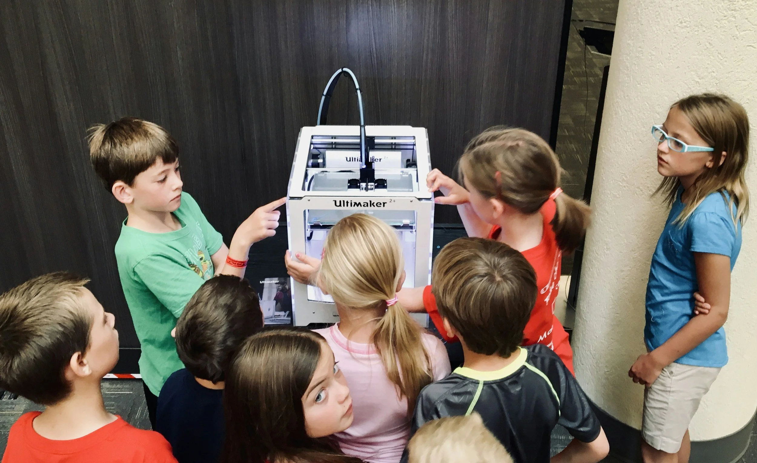 A group of children gathered around a 3D printer, watching it as it operates. Some children are pointing at the printer, and a girl wearing glasses in a blue shirt stands to the right, observing.