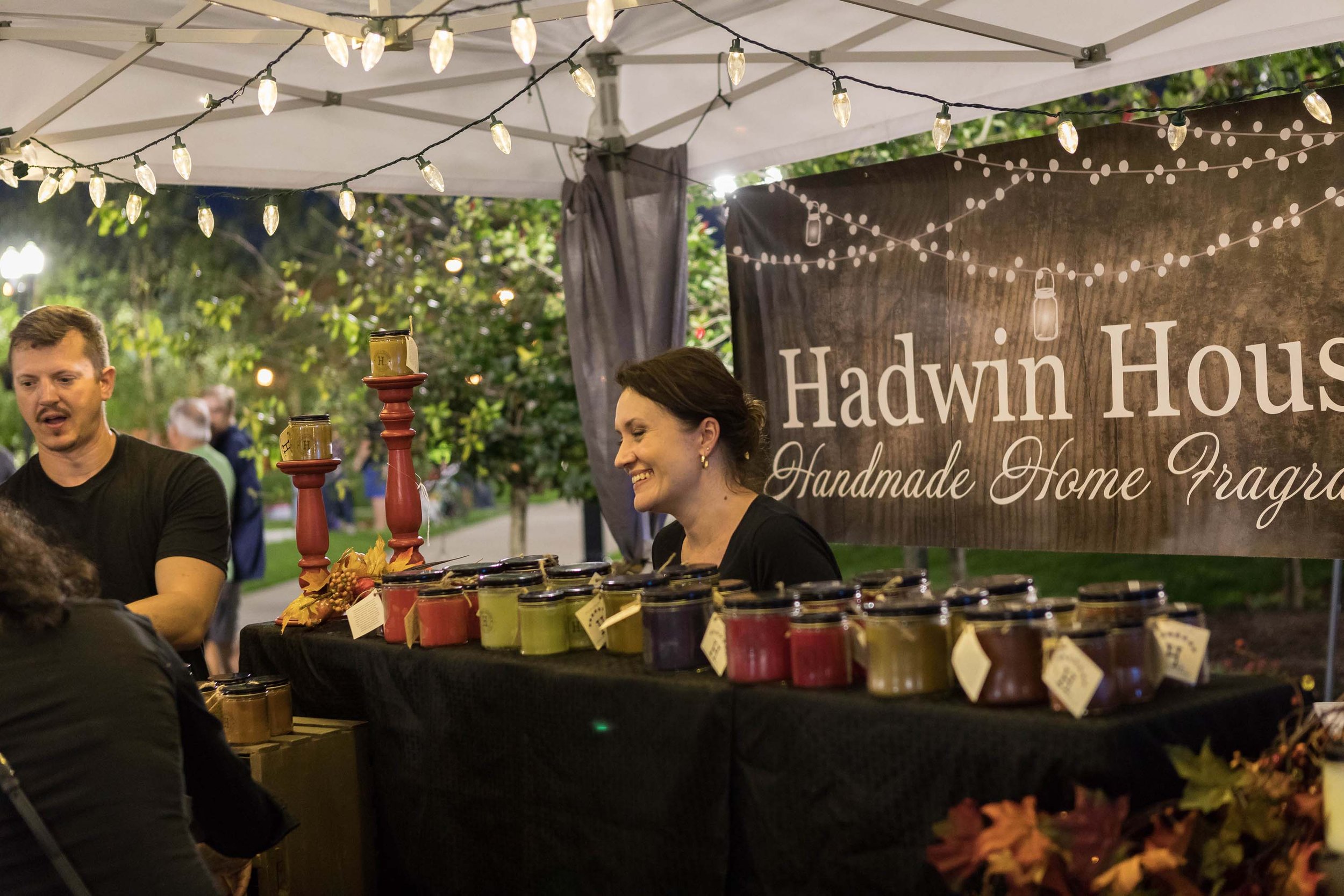 A vendor booth at an outdoor market with illuminated string lights, a large sign reading 'Hawkin House' with the tagline 'Handmade Home Fragrances,' and various jars of products on a black tablecloth, with people in the background and greenery around.