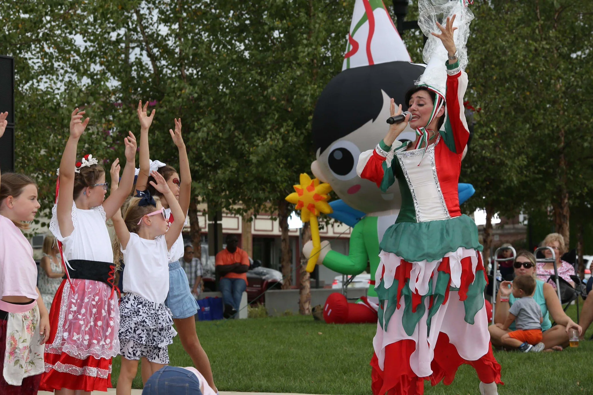Children interacting with a clown performer at an outdoor event, with a large cartoon character statue in the background and people sitting and standing around.