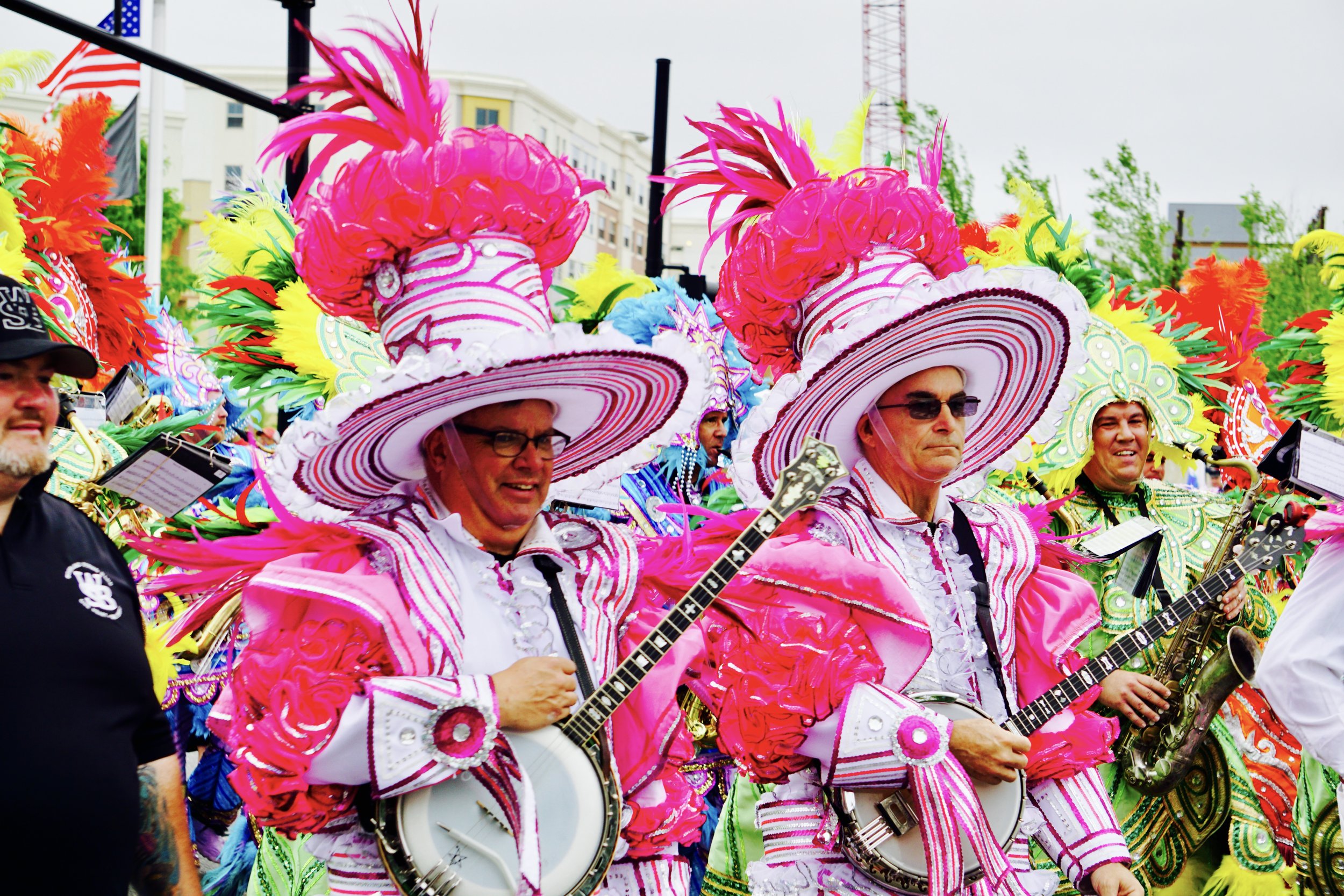 Colorful parade participants wearing large, elaborate pink hats with feathers and dresses, some playing guitars and drums, during an outdoor festival.