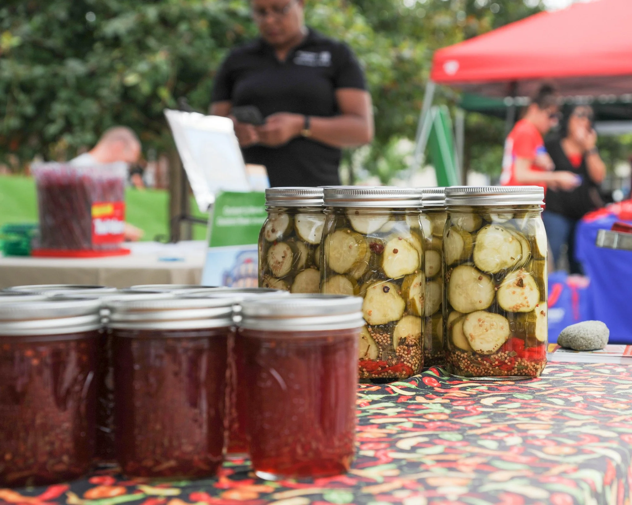Jars of pickled vegetables on display at an outdoor market with people in the background.