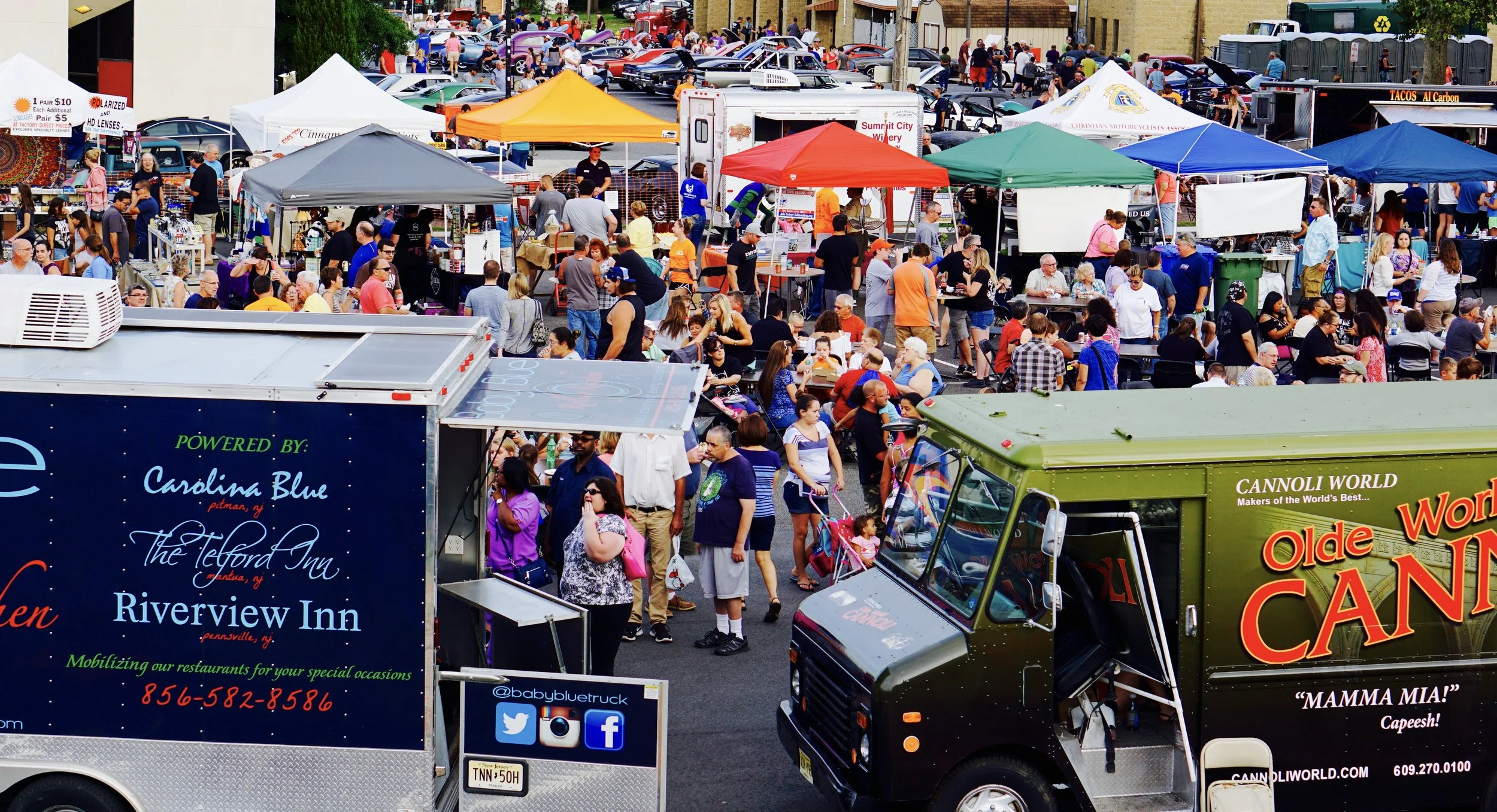 Crowd gathered at a street fair with vendor tents, food trucks, and vintage cars displayed in the background. People are walking, sitting, and socializing.