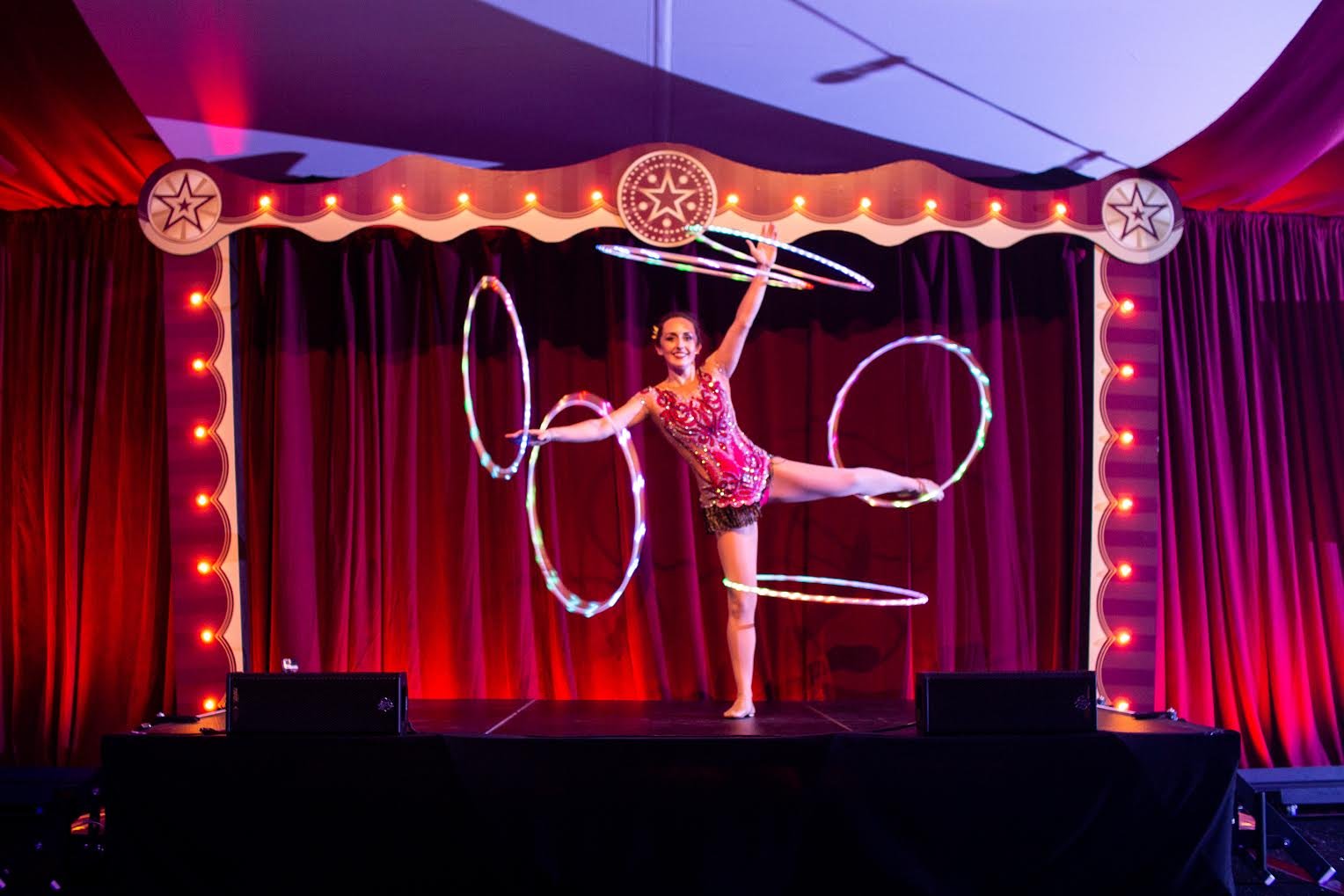 A female hula hoop performer on stage in a red sequined costume, spinning multiple illuminated hula hoops during a circus or show.