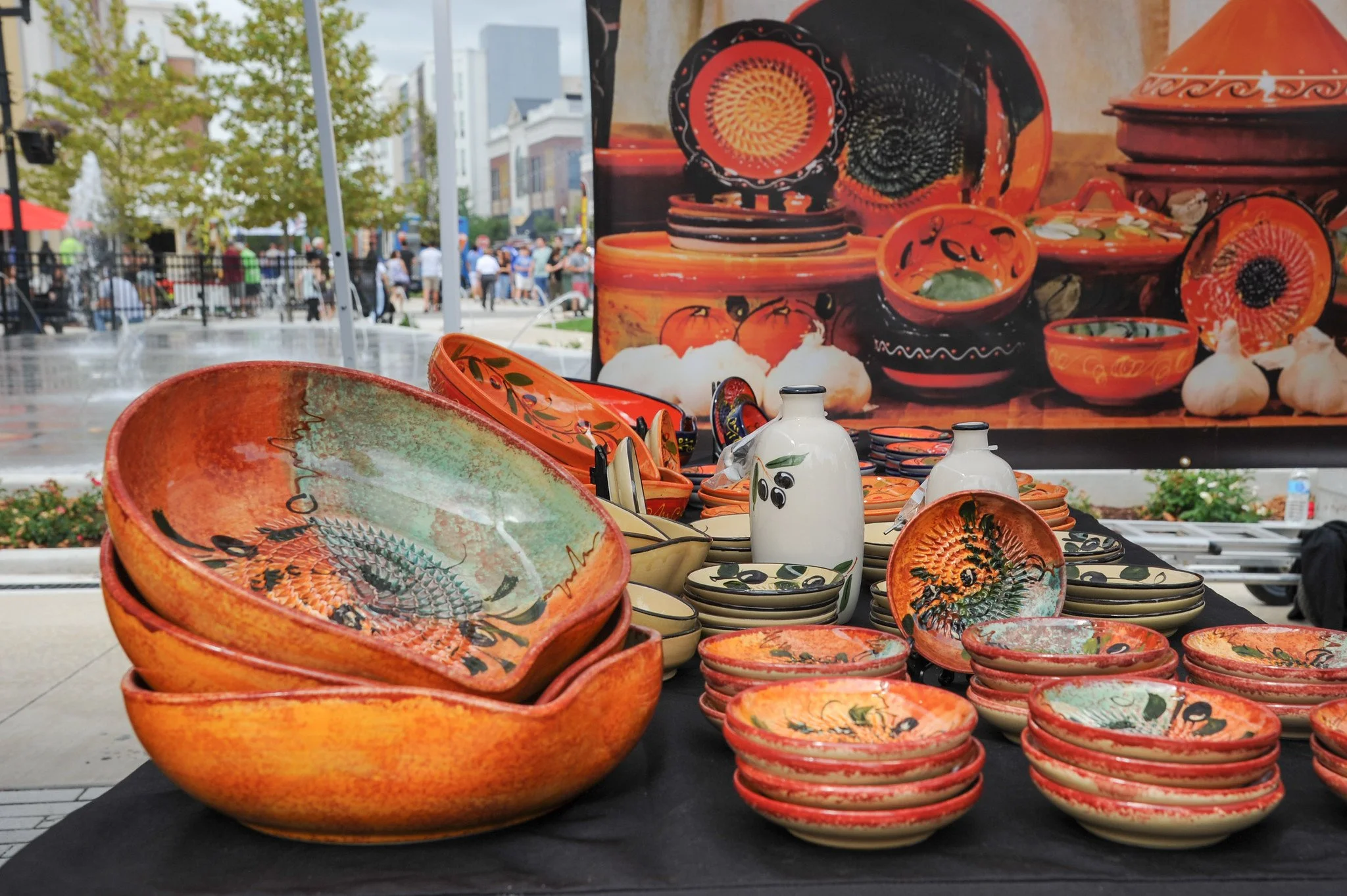 Colorful ceramic pottery on display at an outdoor market, with a painted backdrop and people walking in the background.