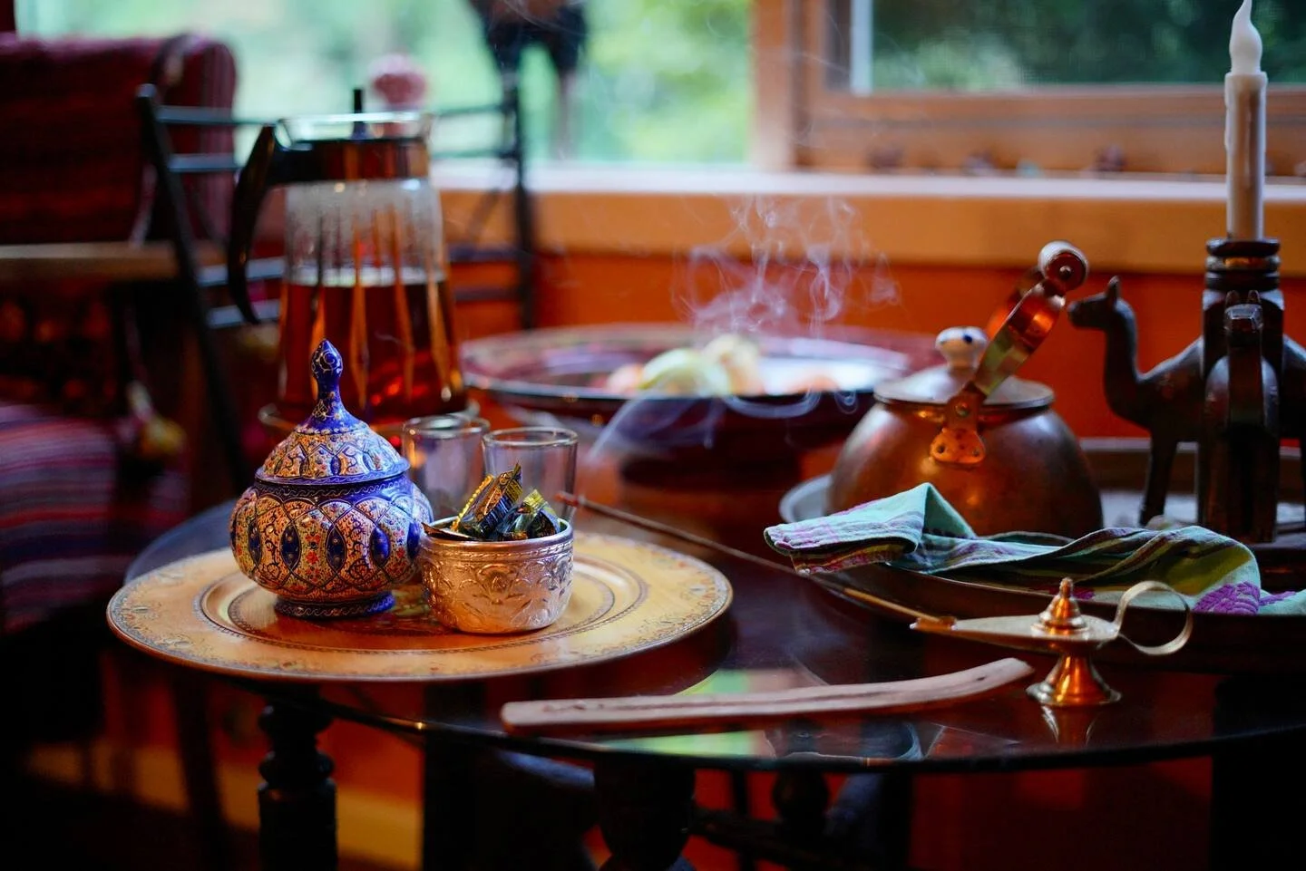 A table set with traditional Middle Eastern items, including a decorative teapot, a copper cezve pot with steam, small glasses, and an ornate tray, with a bowl of food in the background, near a window with greenery outside.
