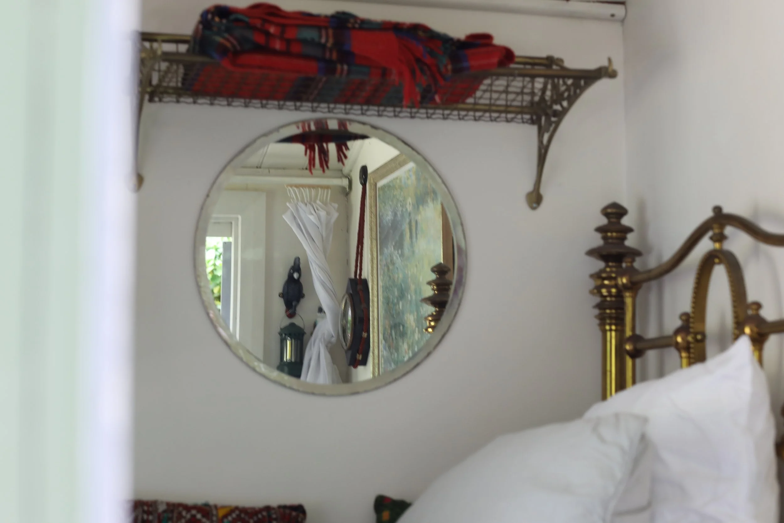 A bedroom with a white wall, a round mirror, a wooden bed with brass posts, and a colorful bedspread.