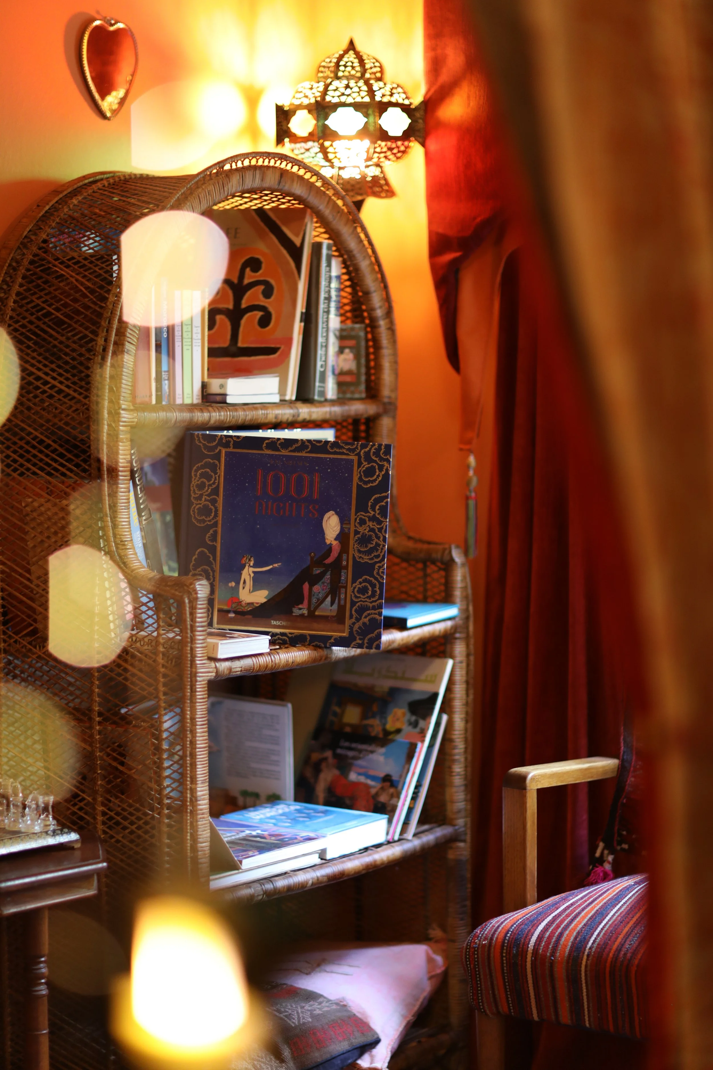A wooden bookshelf filled with books, a table with books and papers, a striped upholstered chair, a decorative lamp on the wall emitting warm light, and a wall mirror reflecting part of the room, all in a cozy, warmly lit room.