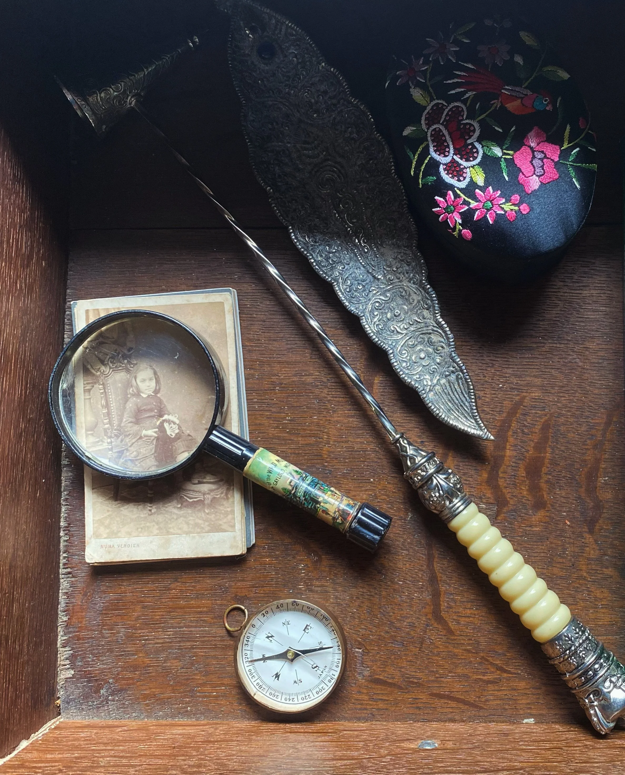 An antique incense holder, a deck of vintage photographs, a magnifying glass, a compass, a colorful embroidered box, and a silver and ivory candle snuffer are arranged in a wooden drawer.