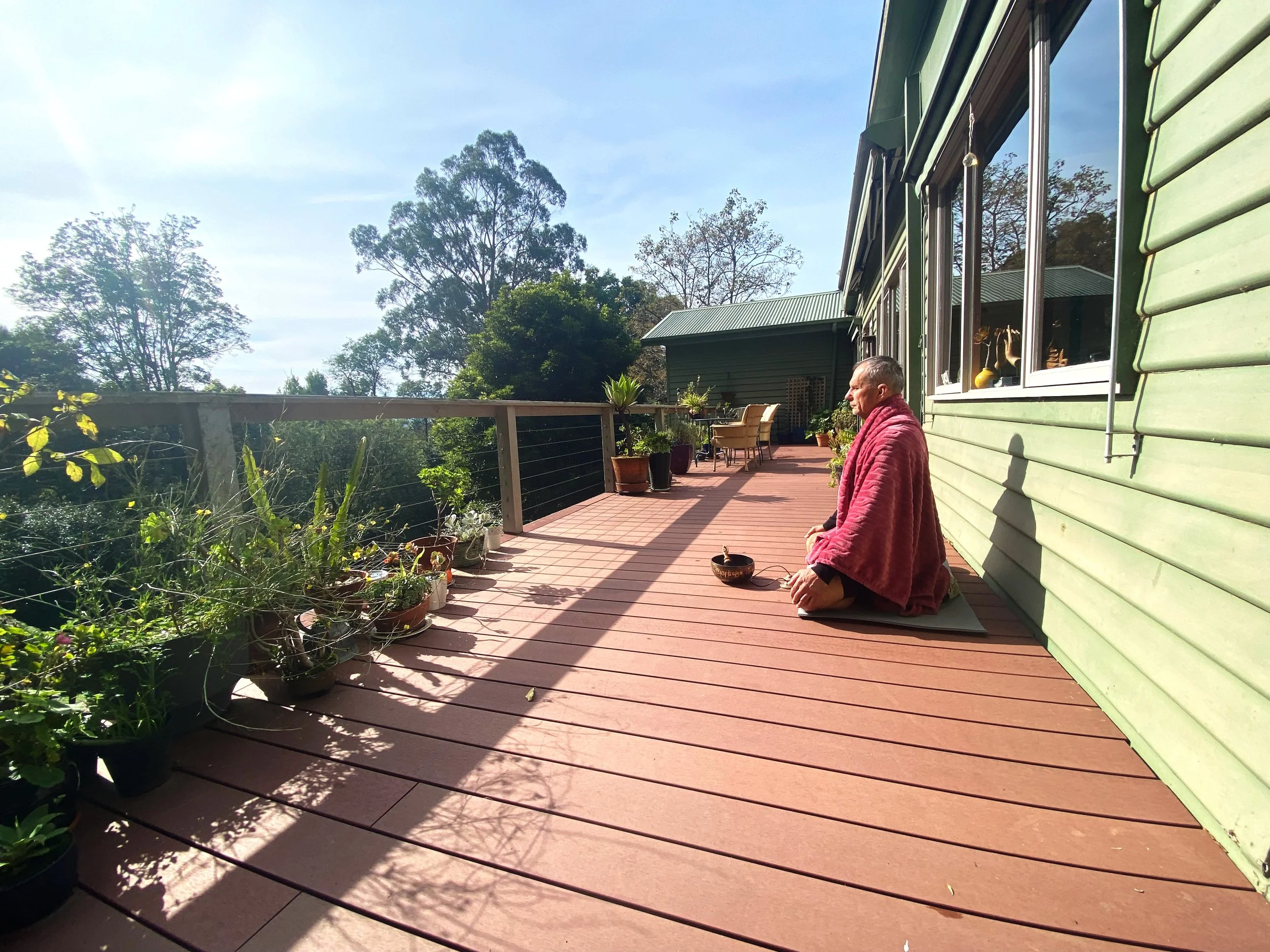A man kneeling on a wooden deck outside a house, practicing meditation with eyes closed, wearing a coral pink wrap. The deck has potted plants and is bordered by a metal railing with trees and a blue sky in the background.