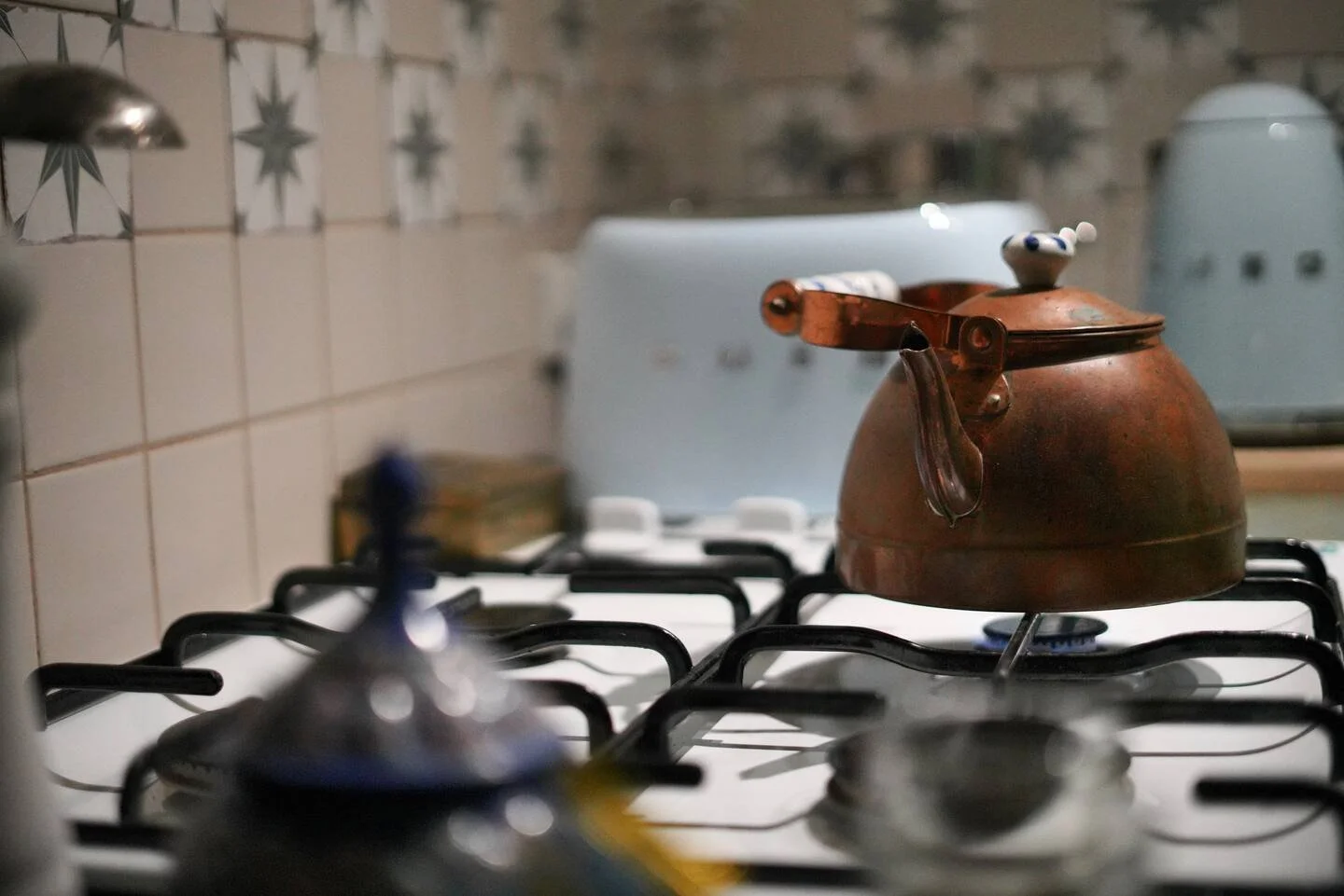 A vintage copper kettle on a stovetop burner with a ceramic lid handle, in a kitchen setting with tiled walls and a white appliance in the background.