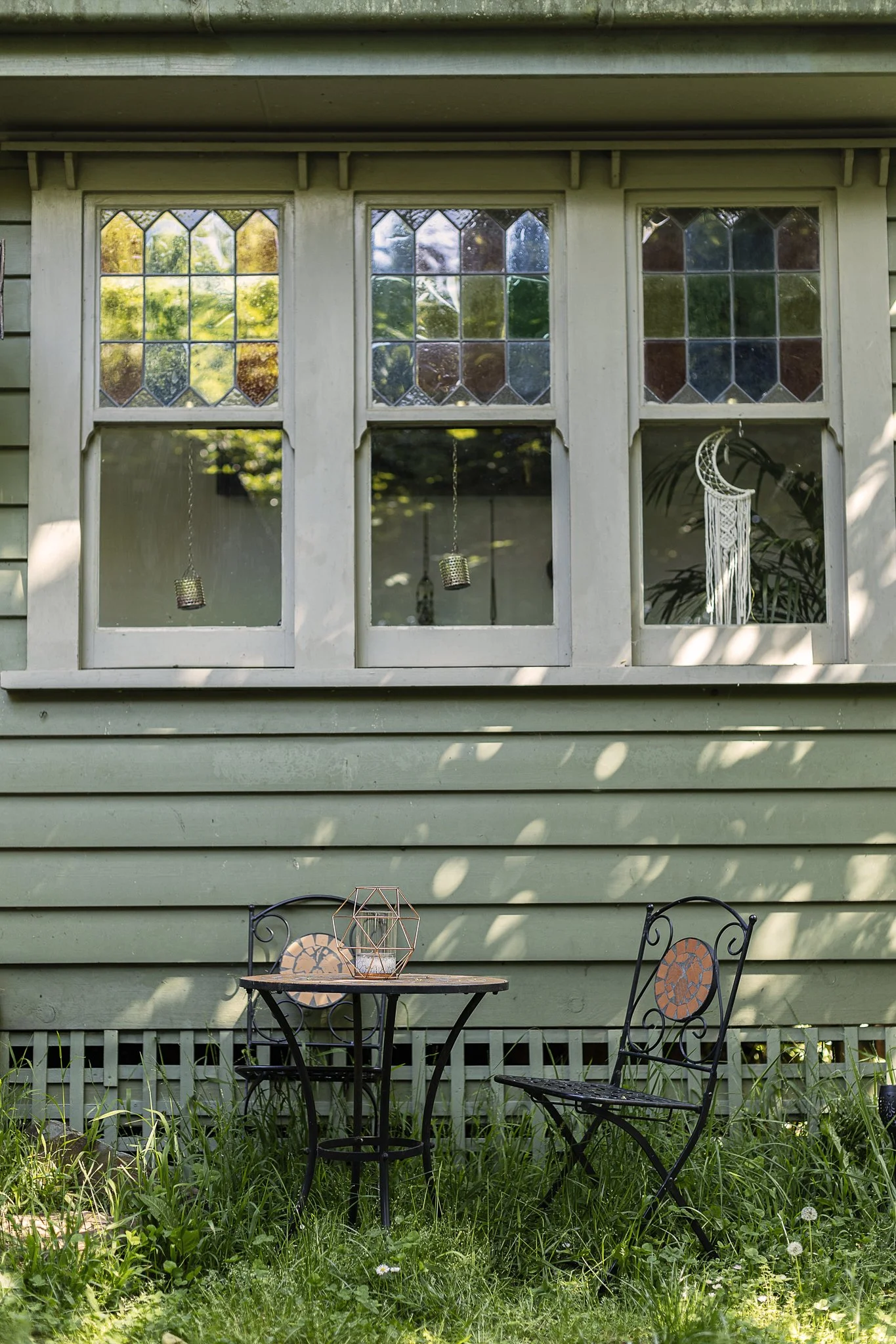 A green house with three large windows, a table, and two chairs outside in a garden. The windows have decorative stained glass, and there's a wind chime and a hanging decoration visible inside the window. Grass and plants surround the seating area.