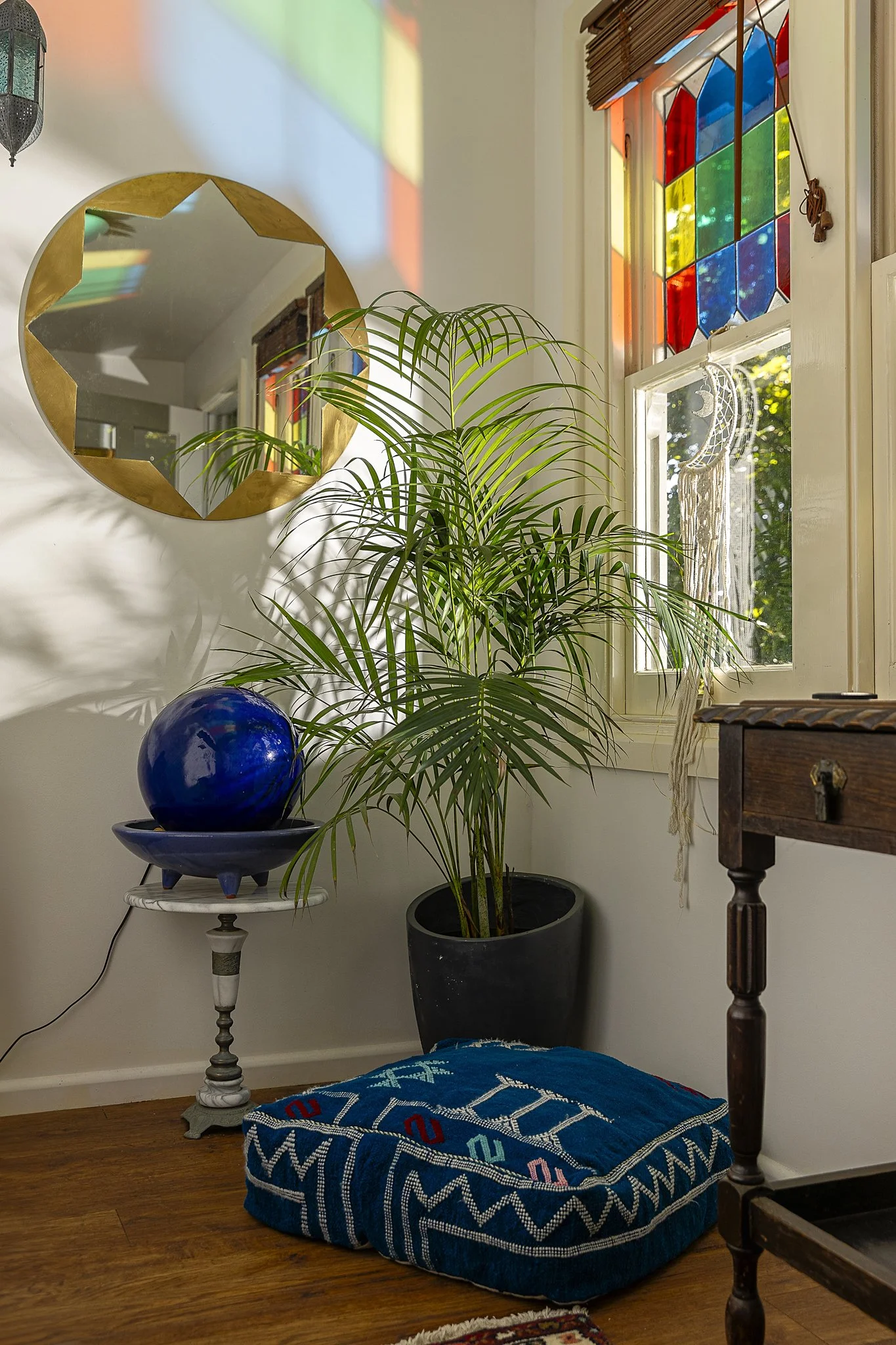 A cozy home interior with a large potted plant near a window with colorful stained glass. A mirror with a gold frame reflects the room, while a blue decorative sphere sits on a white side table. There is a dark wooden table on the right, a blue patte