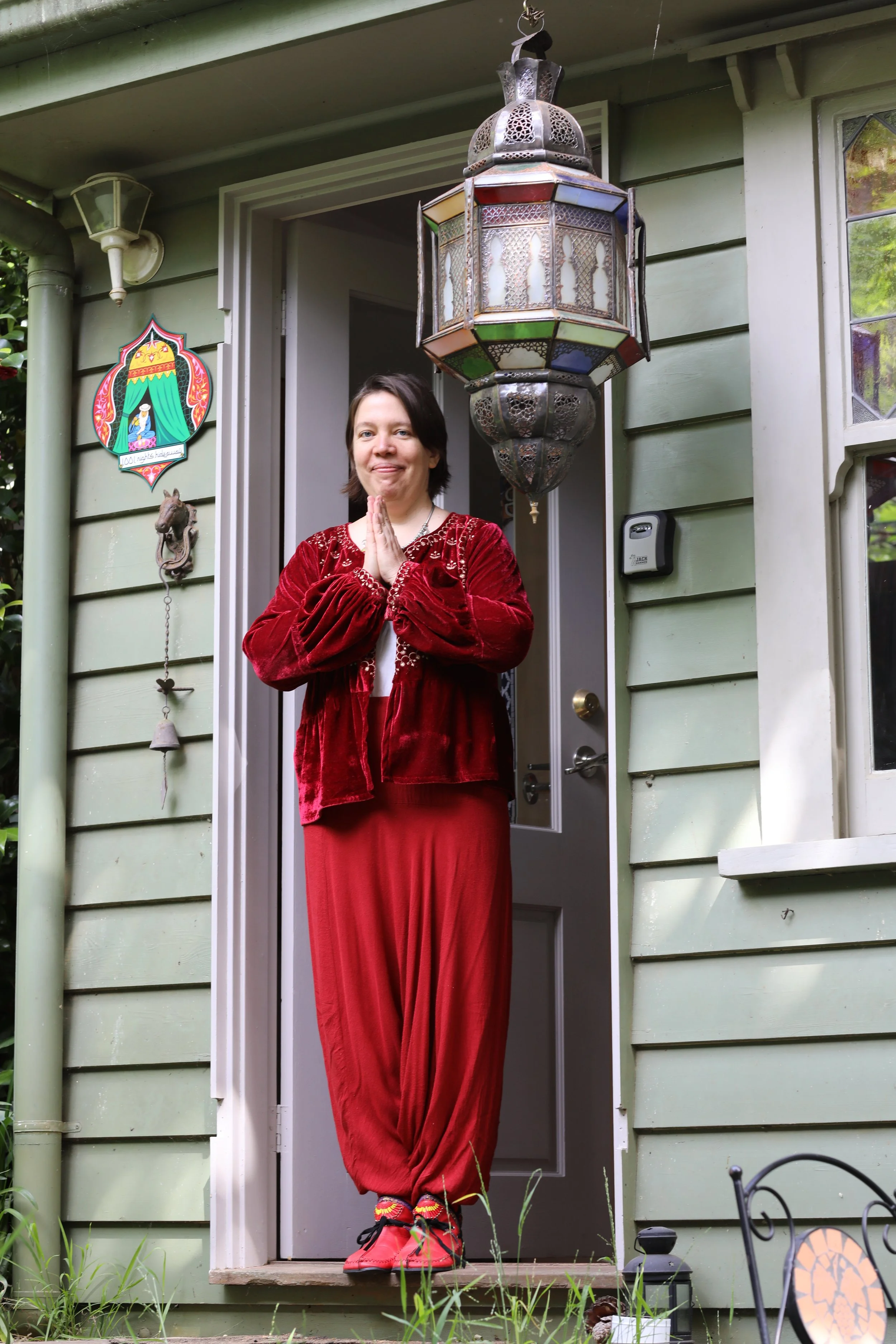 A woman standing at a porch doorway with her hands pressed together in a prayer gesture. She is wearing red velvet clothing and bright red shoes, and the house is painted green with decorative elements including a bell and a colorful lantern hanging above her.