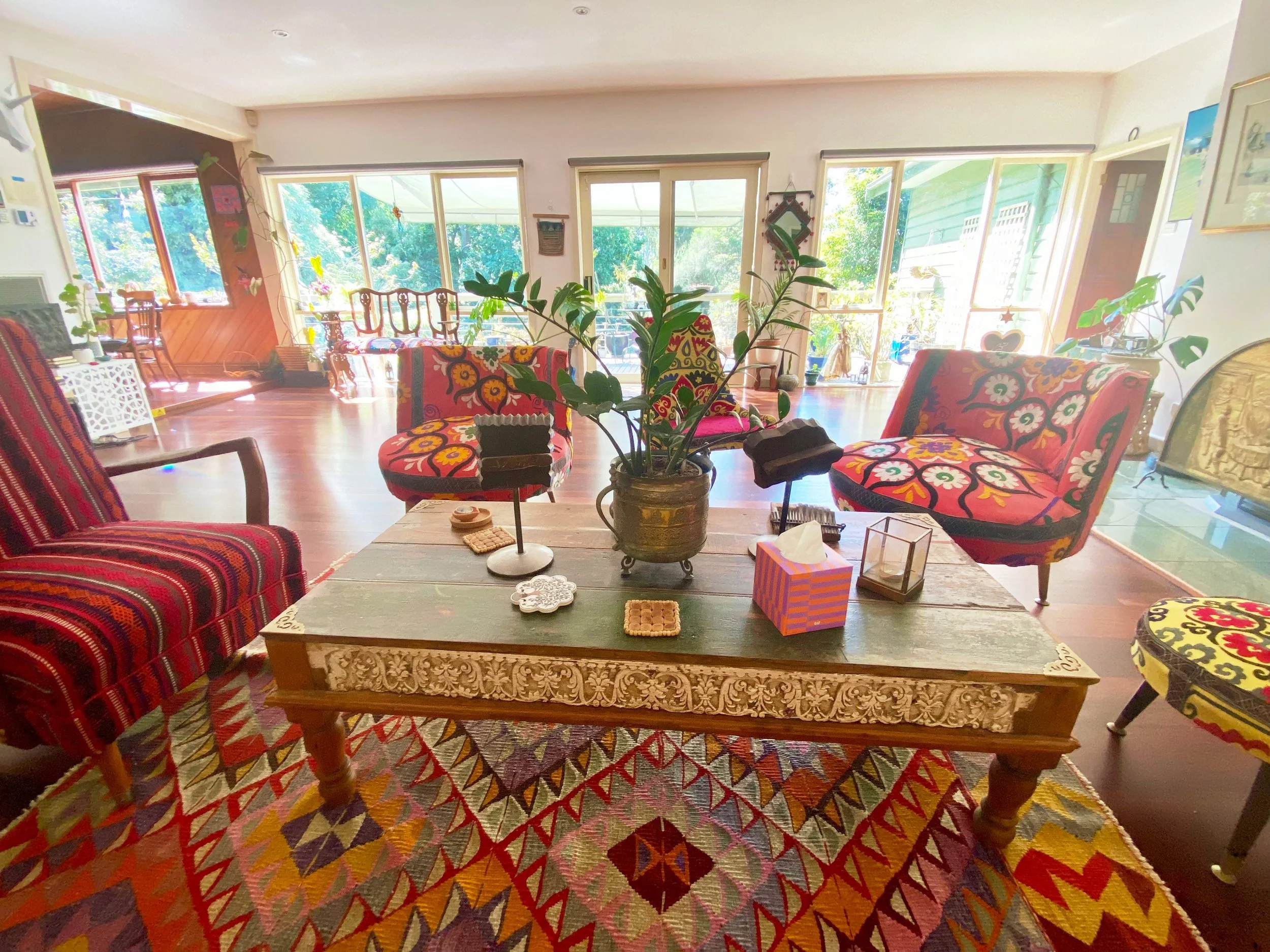 A cozy living room with large windows overlooking trees, decorated with colorful floral-patterned chairs, a wooden coffee table, and various plants and ornaments.