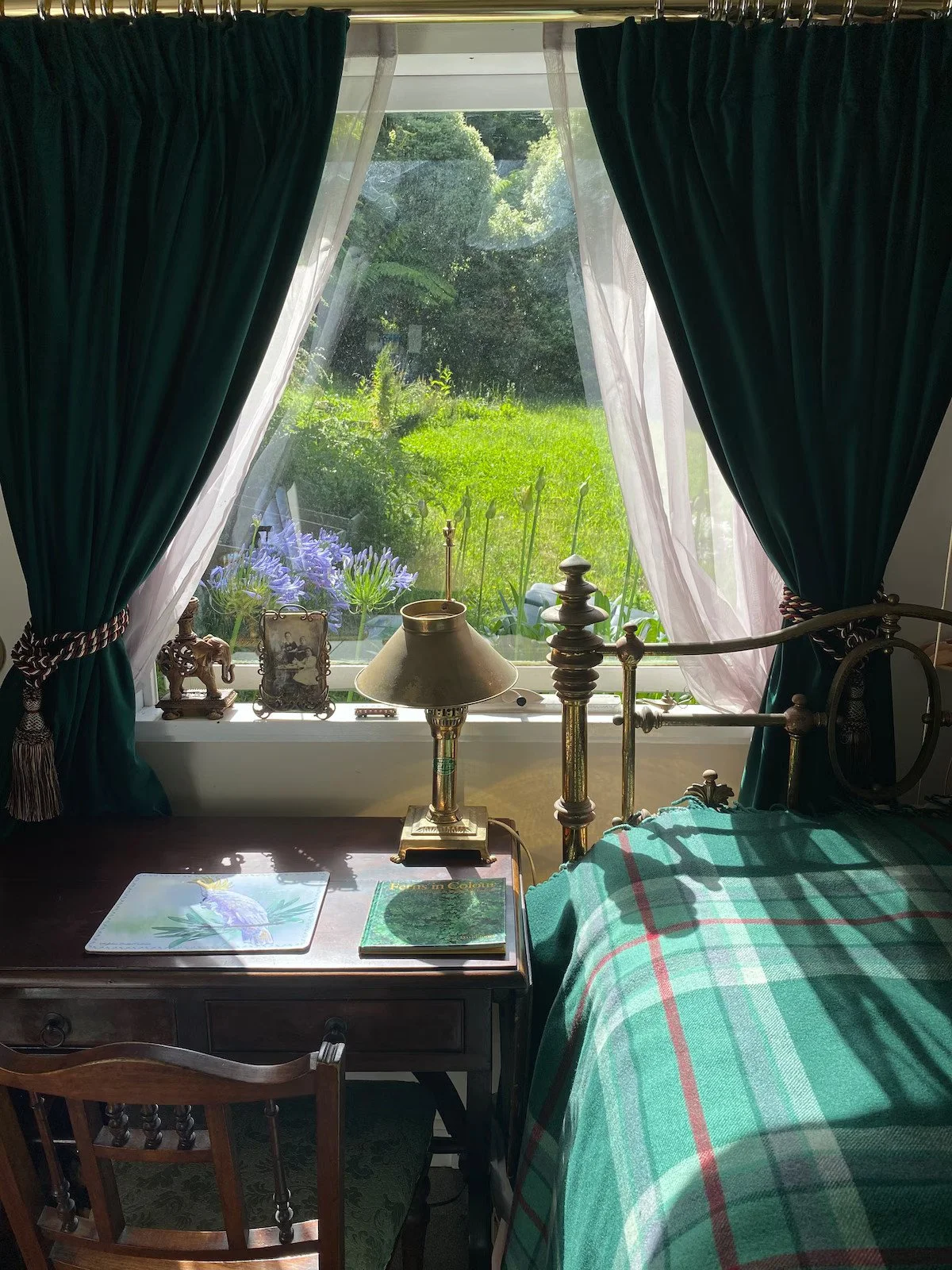 A bedroom with a large window showing a lush green garden outside. The window has dark green curtains tied back with decorative cords and sheer white curtains. On the windowsill, there are small decorative items, including a miniature elephant, a pho