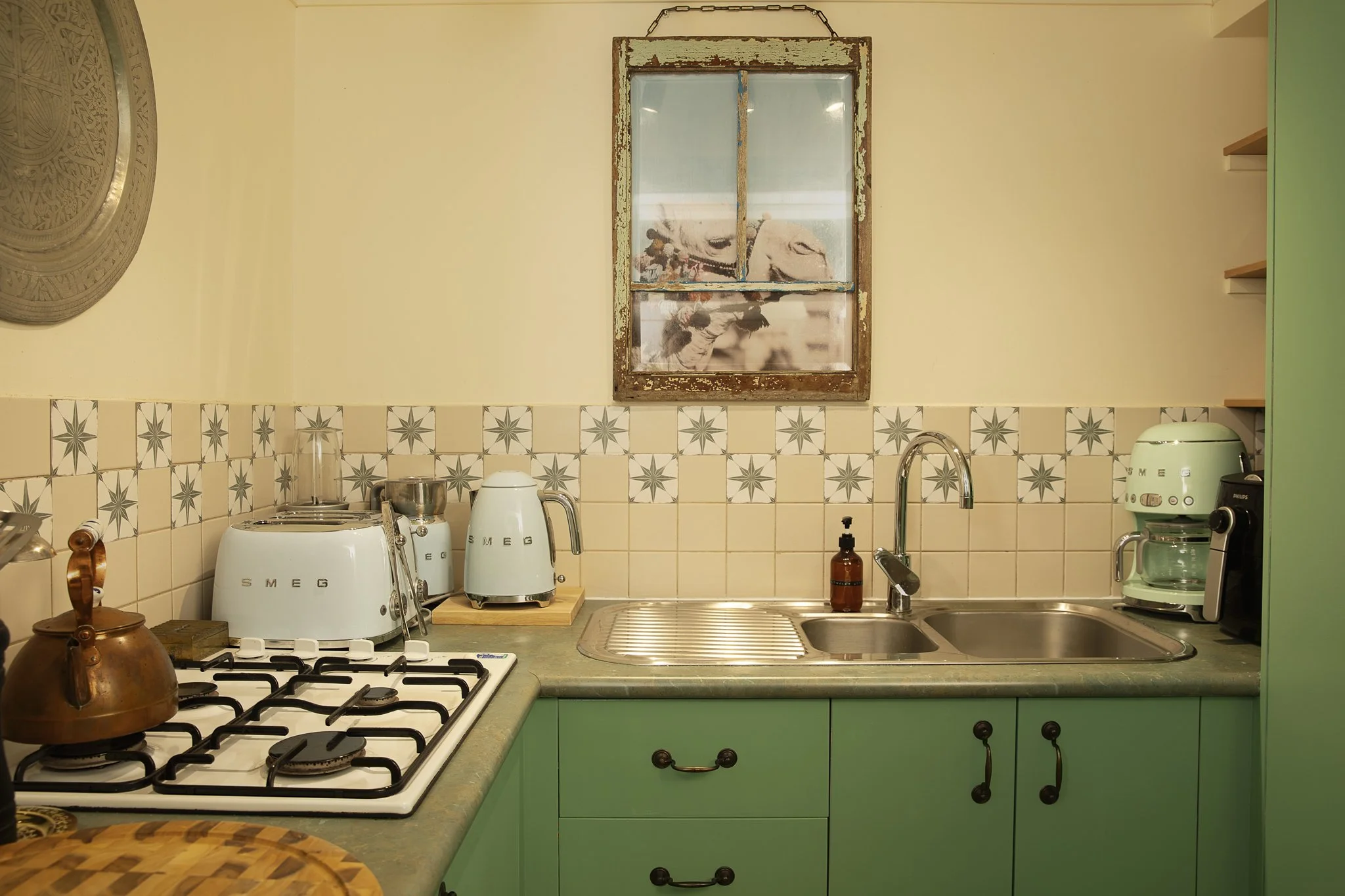 A kitchen with green cabinets, a stainless steel sink, a stovetop, and various appliances like toasters and a coffee maker. There's a small kitchen window with a rustic wooden frame. The walls are painted a light color, partially covered with patterned tiles. Stylish items such as a copper kettle and a wooden cutting board are on the counter.