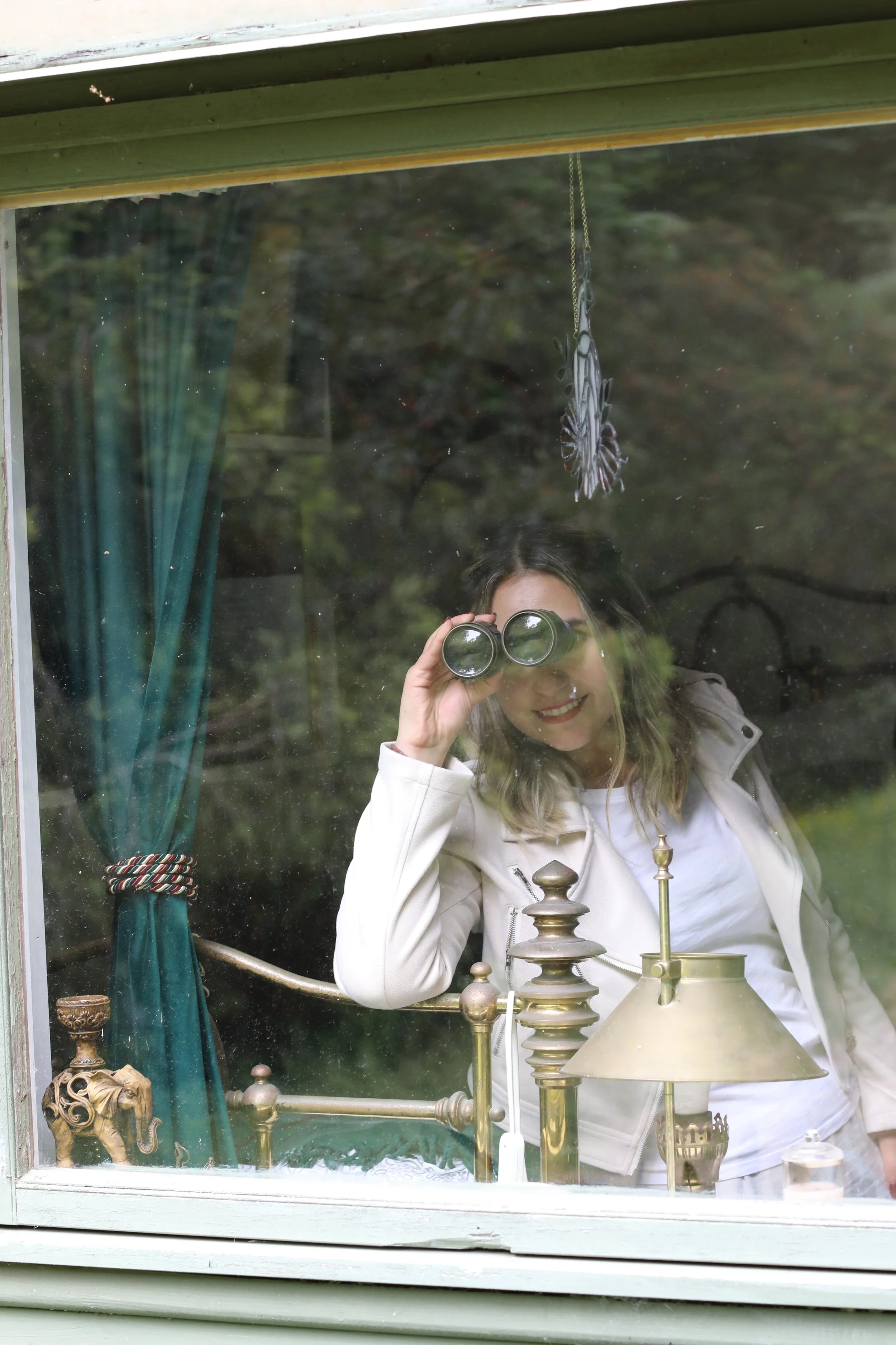 Person with blonde hair and glasses looking through binoculars, standing behind a display of brass and gold-colored objects, inside a window with a green curtain at the side.