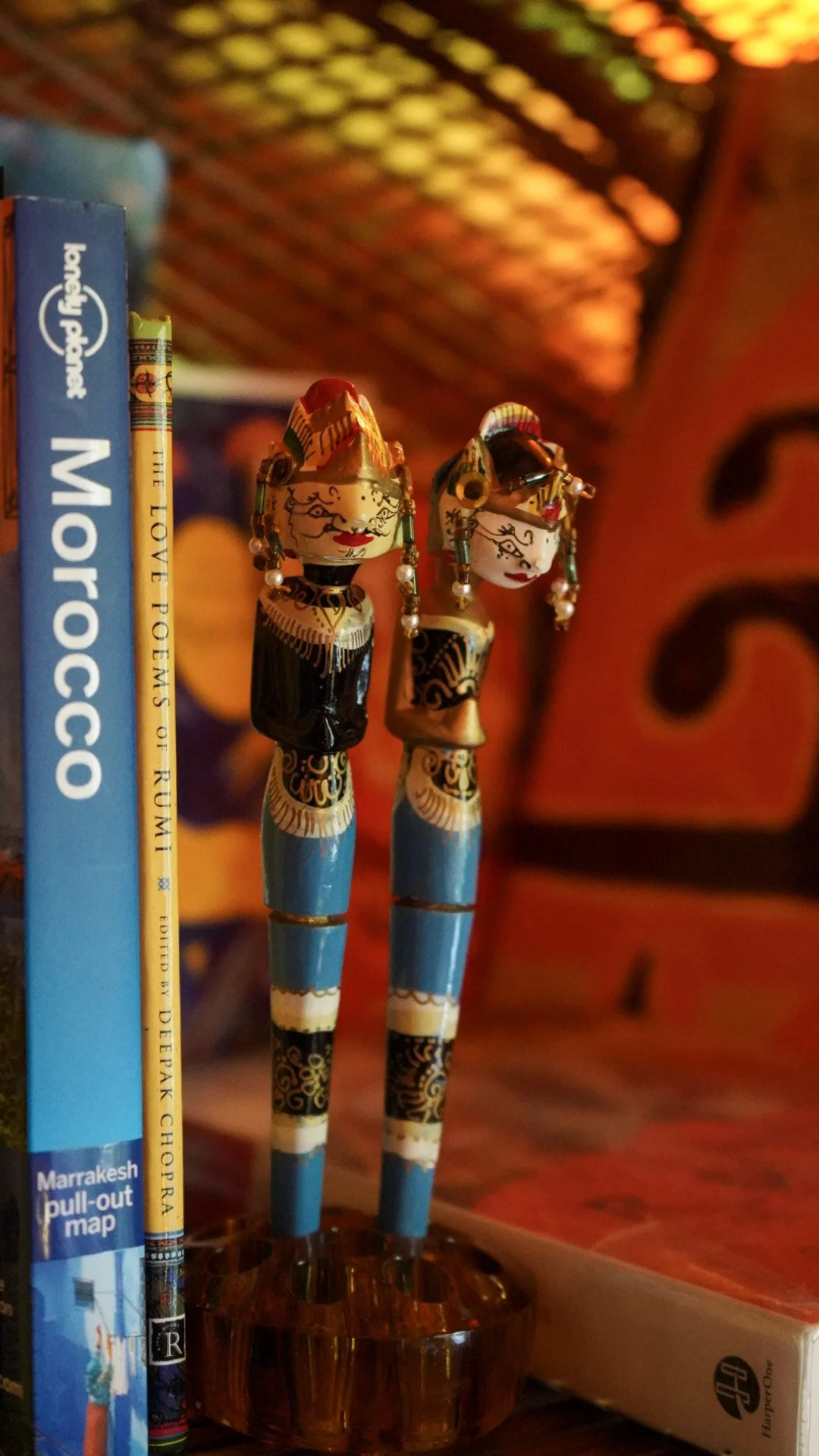 Decorative figurines of two miniature wayang golek characters with detailed faces and traditional attire, standing next to books, in a warm, colorful setting.