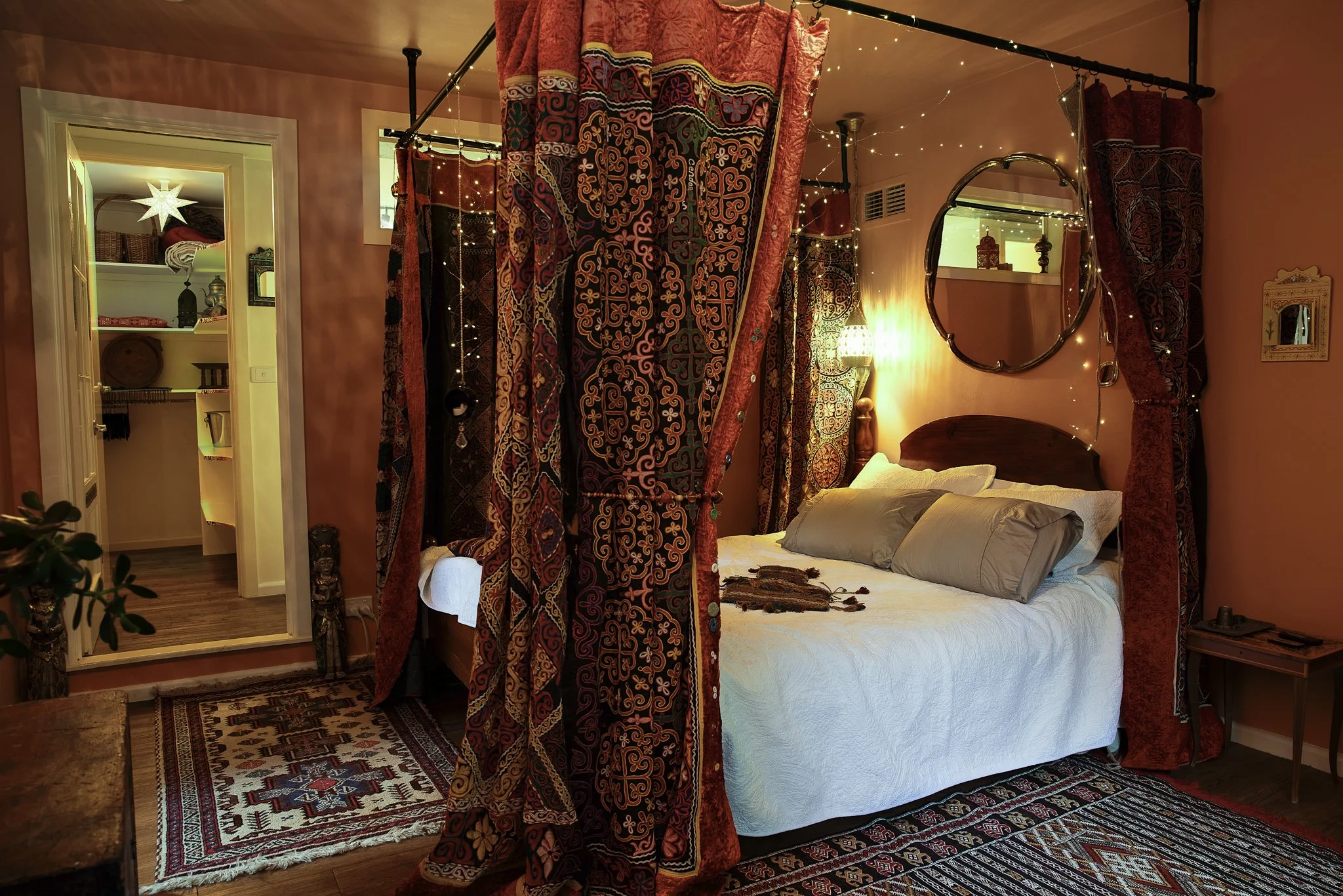 A cozy bedroom with a four-poster bed draped with patterned velvet curtains, with fairy lights hanging on the bedposts. The bed has white bedding and pillows, and there is a small wooden nightstand. A large oval mirror hangs on the wall, reflecting a