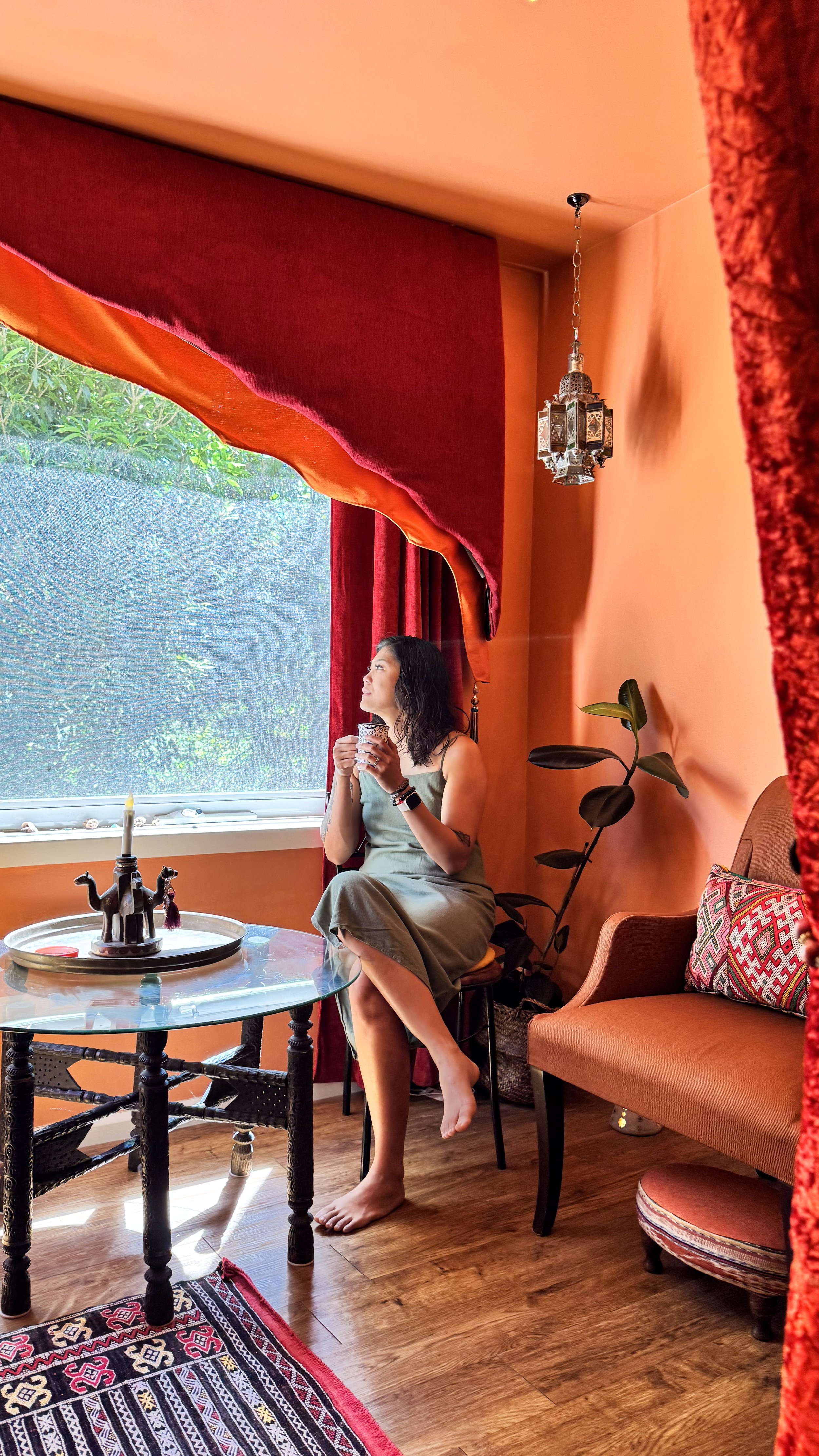 Woman sitting on a chair near a window, holding a cup, with a colorful room interior including orange walls, red curtains, a decorative pillow, a plant, a chandelier, and a table with decor items.