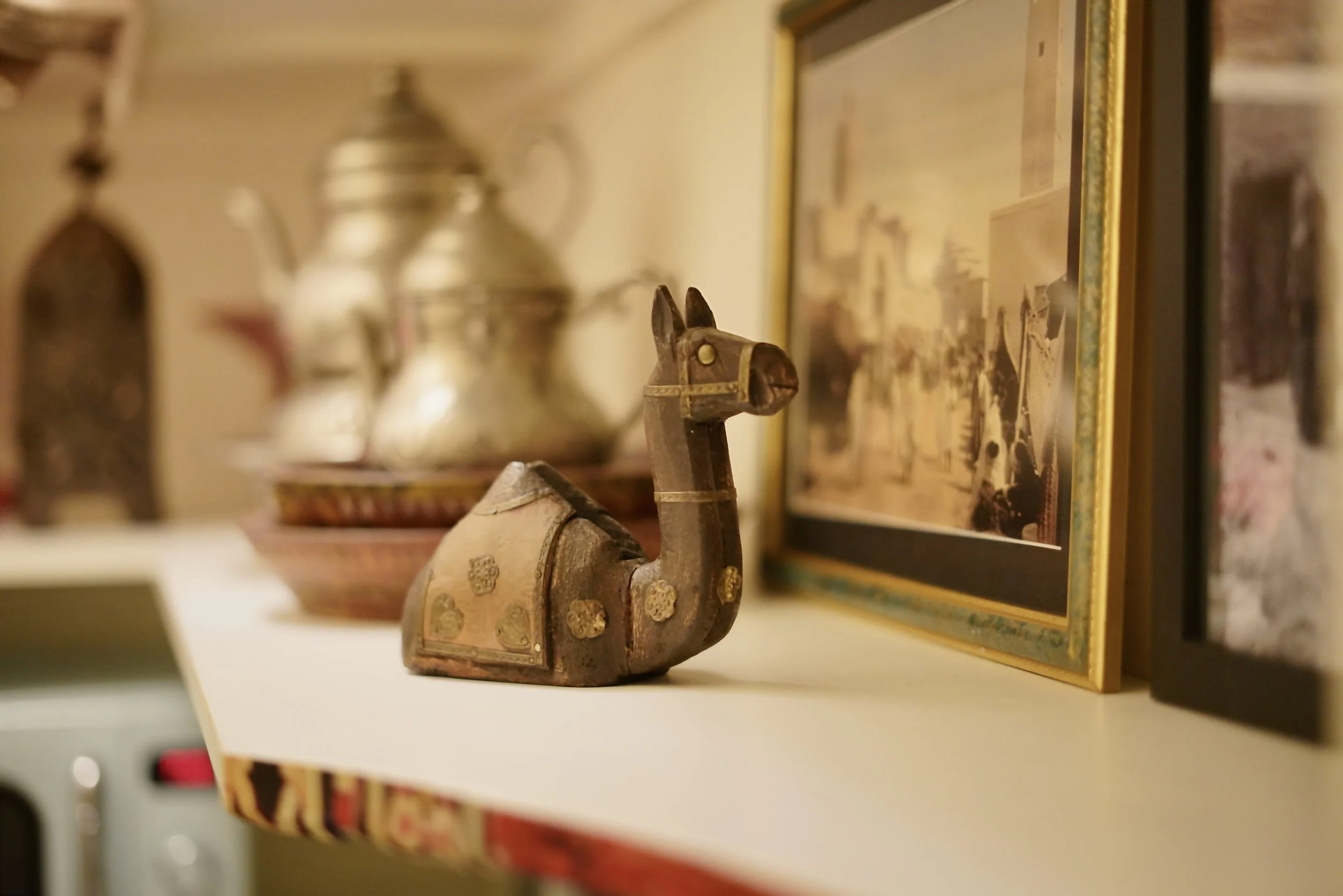 Decorative camel figurine with intricate details, placed on a white shelf, with a framed picture and blurred other objects in the background.