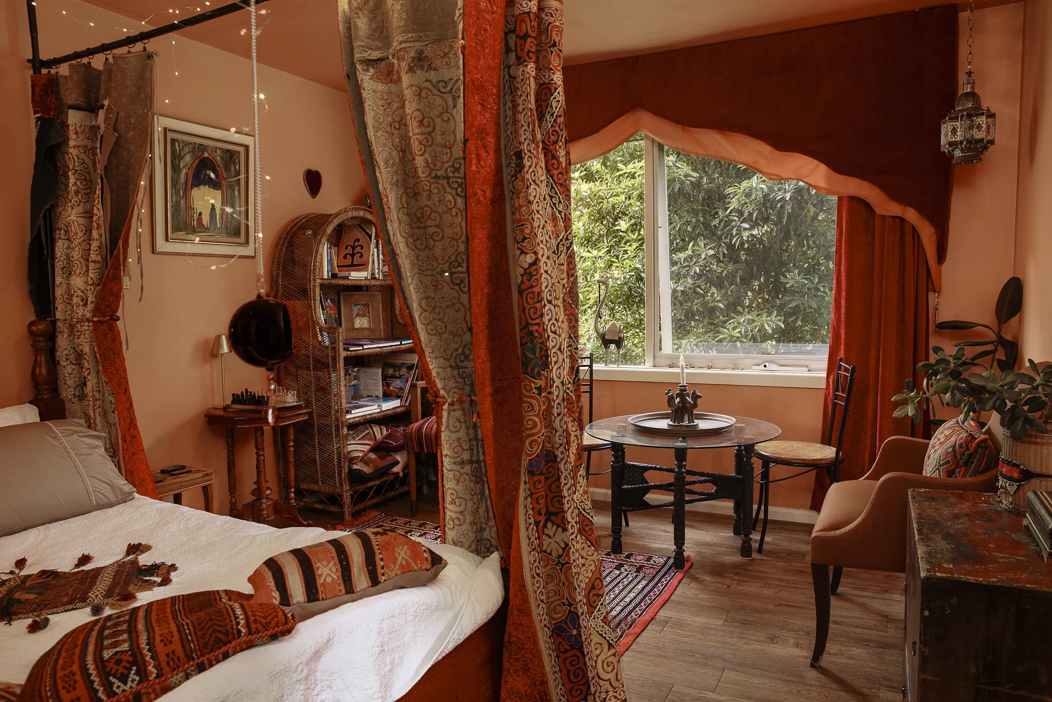 A cozy living room with peach-colored walls and large window with pinkish-orange curtains, decorated with tapestry and artwork, featuring a round glass dining table, cushioned chairs, and a bookshelf with plants and decorative items.