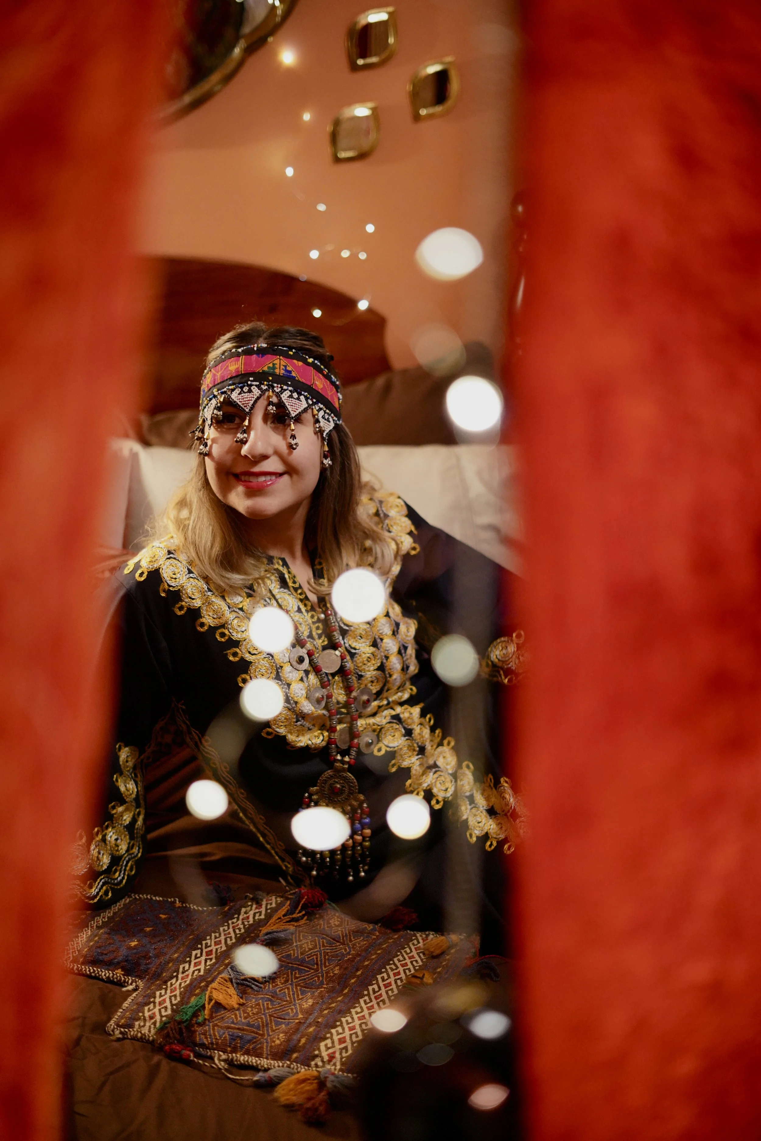 A woman with medium-length blonde hair, wearing traditional tribal attire, including a beaded headdress, earrings, necklaces, and a embroidered dress, smiling, with the photo taken through an orange-red fringed frame, creating a festive and cultural 